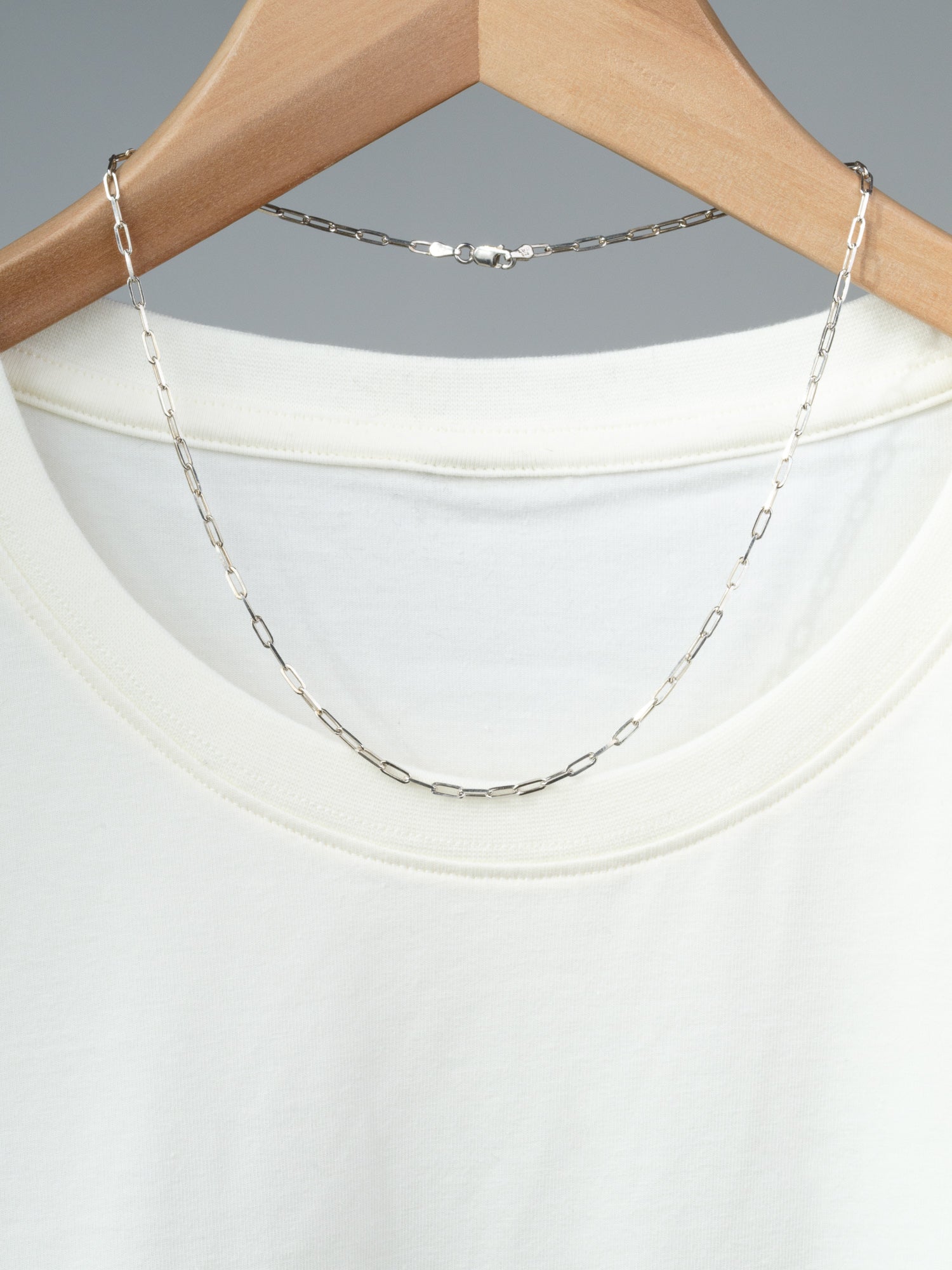 The Kai 80 Paper Clip Necklace by ID Menswear—crafted from thin 925 Sterling Silver with elongated links—rests over the neckline of a plain white T-shirt displayed on a wooden hanger against a neutral gray background.