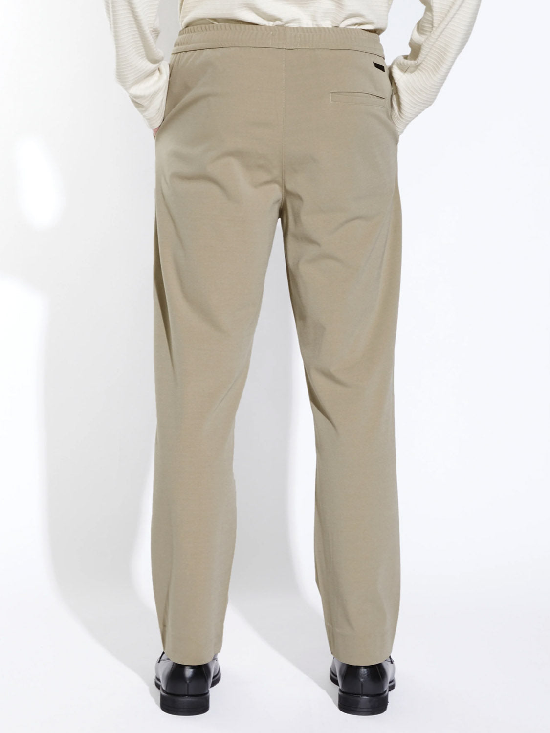 A person is shown from the back against a plain white background, wearing Civil Society's Klien Loose Fit Dressy Trouser in light beige with a tucked-in off-white top and black shoes.