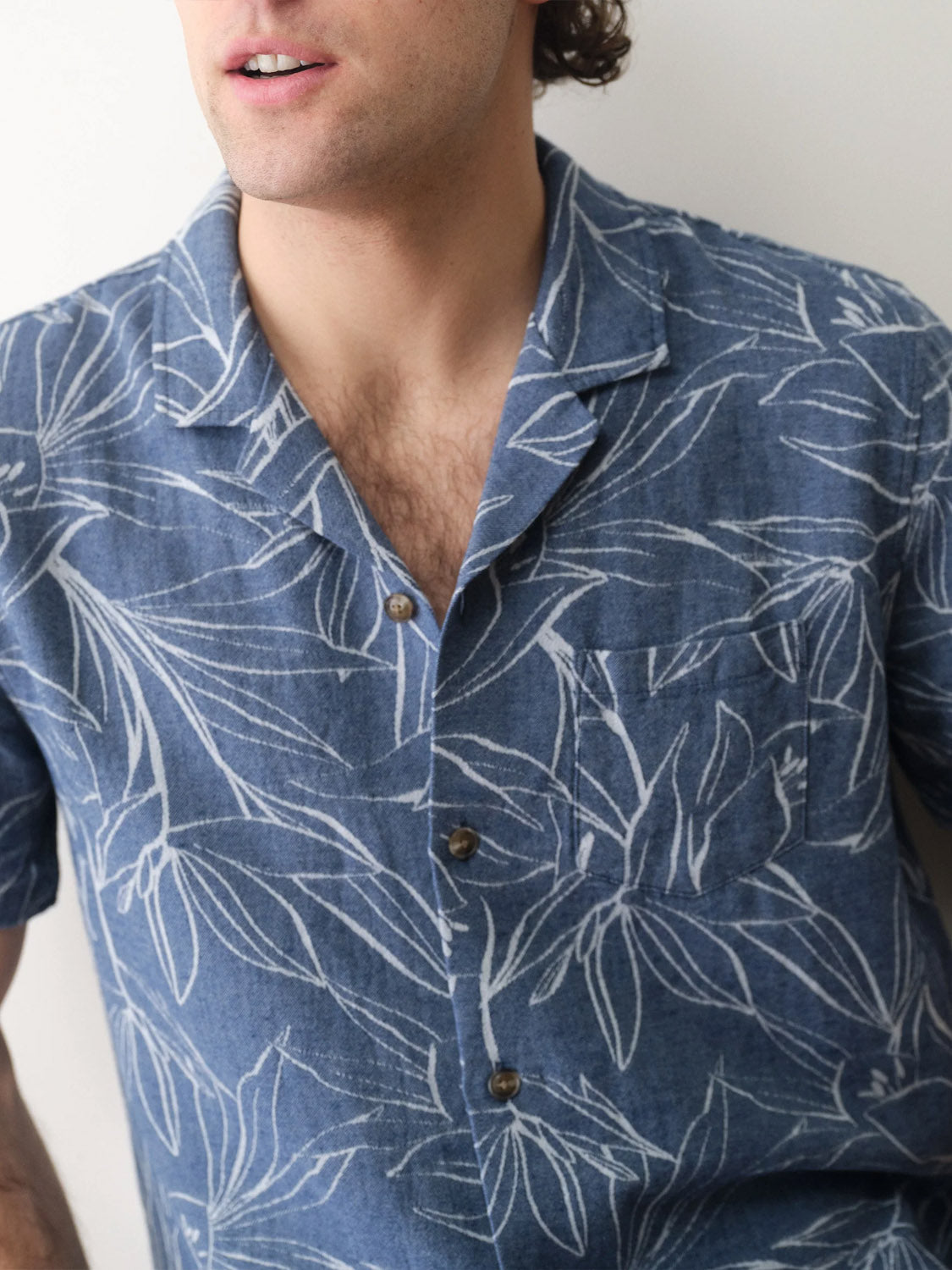 A man wearing the Marine Layer Leo Jacquard Resort Shirt, featuring a blue fabric with a white woven leaf design, front pocket, and brown buttons. Only his lower face and upper torso are visible.