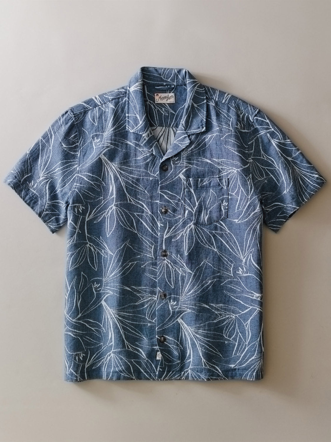 The Leo Jacquard Resort Shirt by Marine Layer is a short-sleeve, button-up with a denim-blue base and white woven leaf pattern, plus a front chest pocket and collar, shown flat on a light-colored surface.