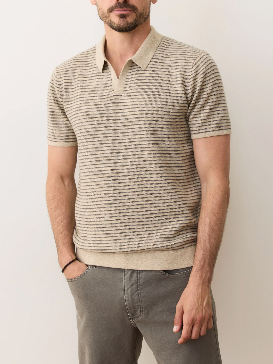 A man wears the Marine Layer Liam Sweater Polo in beige and light brown stripes with gray pants, standing against a plain light background with his hands at his sides.