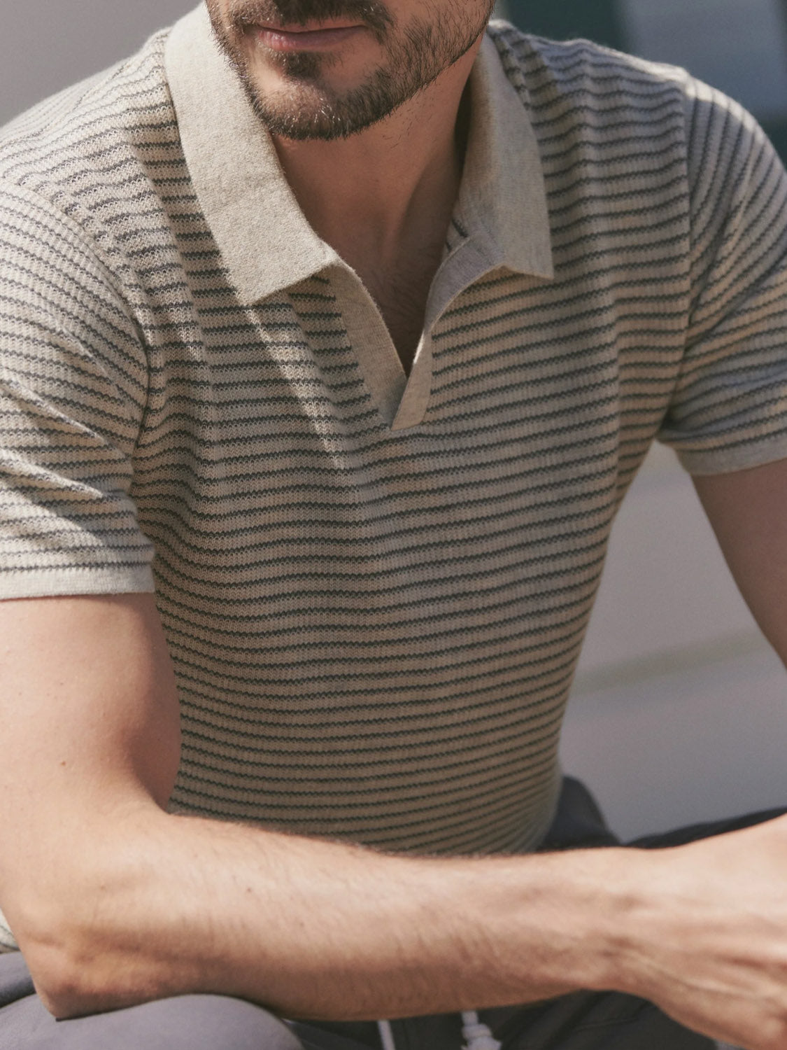 A man with a trimmed beard sits outdoors, wearing the Marine Layer Liam Sweater Polo—a short-sleeved, beige and gray striped sweater polo with a light-colored collar. His face is partially visible.