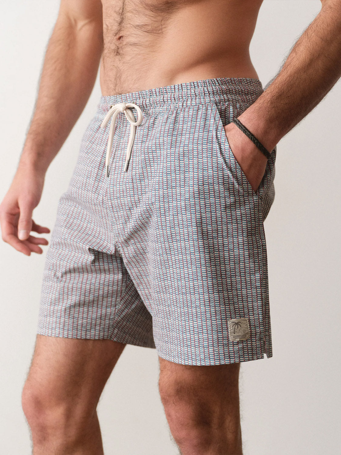 A man stands with one hand in his pocket, wearing Marine Layer Lined Sport and Swim Short 6" featuring a white drawstring and a small patch on the left leg. The light-colored shorts are patterned; image is cropped above the shoulders and below the knees.