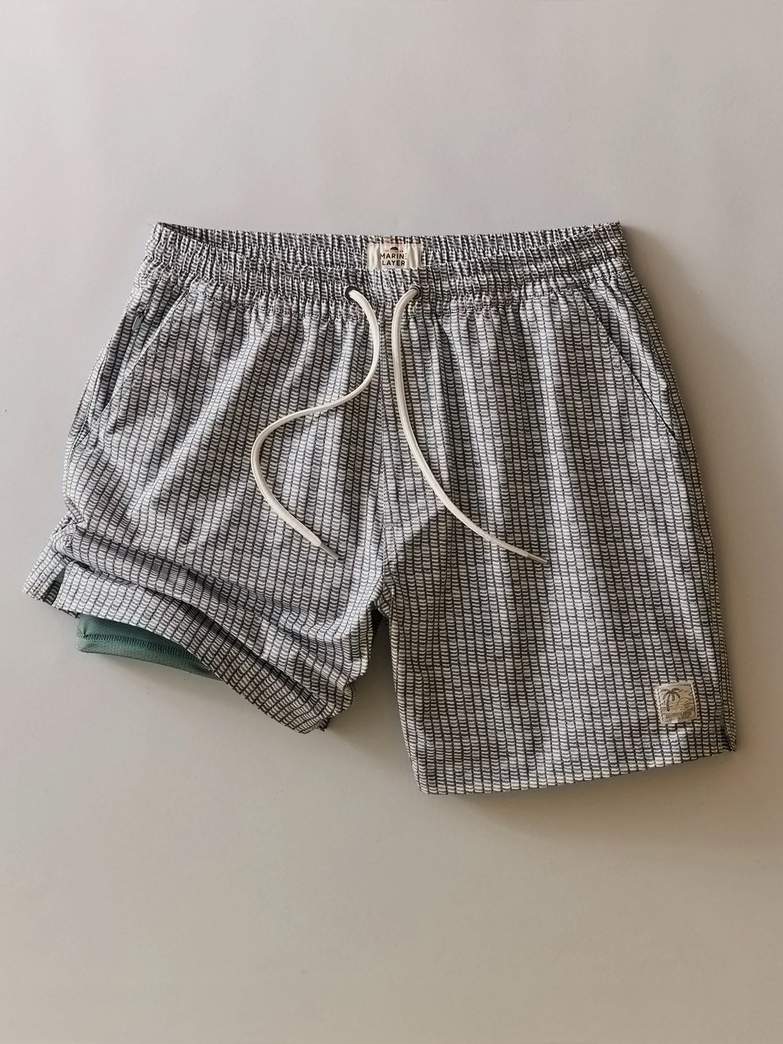 Marine Layer's Lined Sport and Swim Short 6" features gray and white checks, a white drawstring, side pockets, comfy boxer brief liner, light teal accent on the left leg, and quick-dry fabric—perfect for your next getaway.