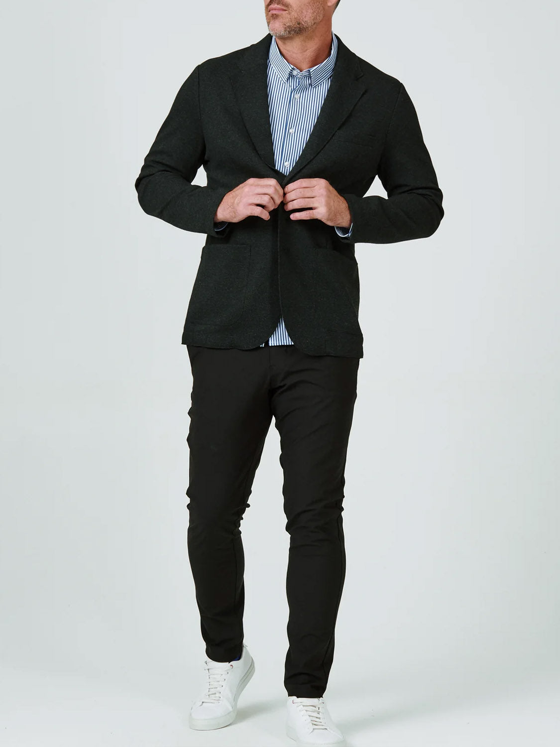 A man, face partially out of frame, adjusts his 7 Diamonds Generation Malibu Blazer over a blue and white striped shirt, paired with black pants and white sneakers, standing against a plain background.