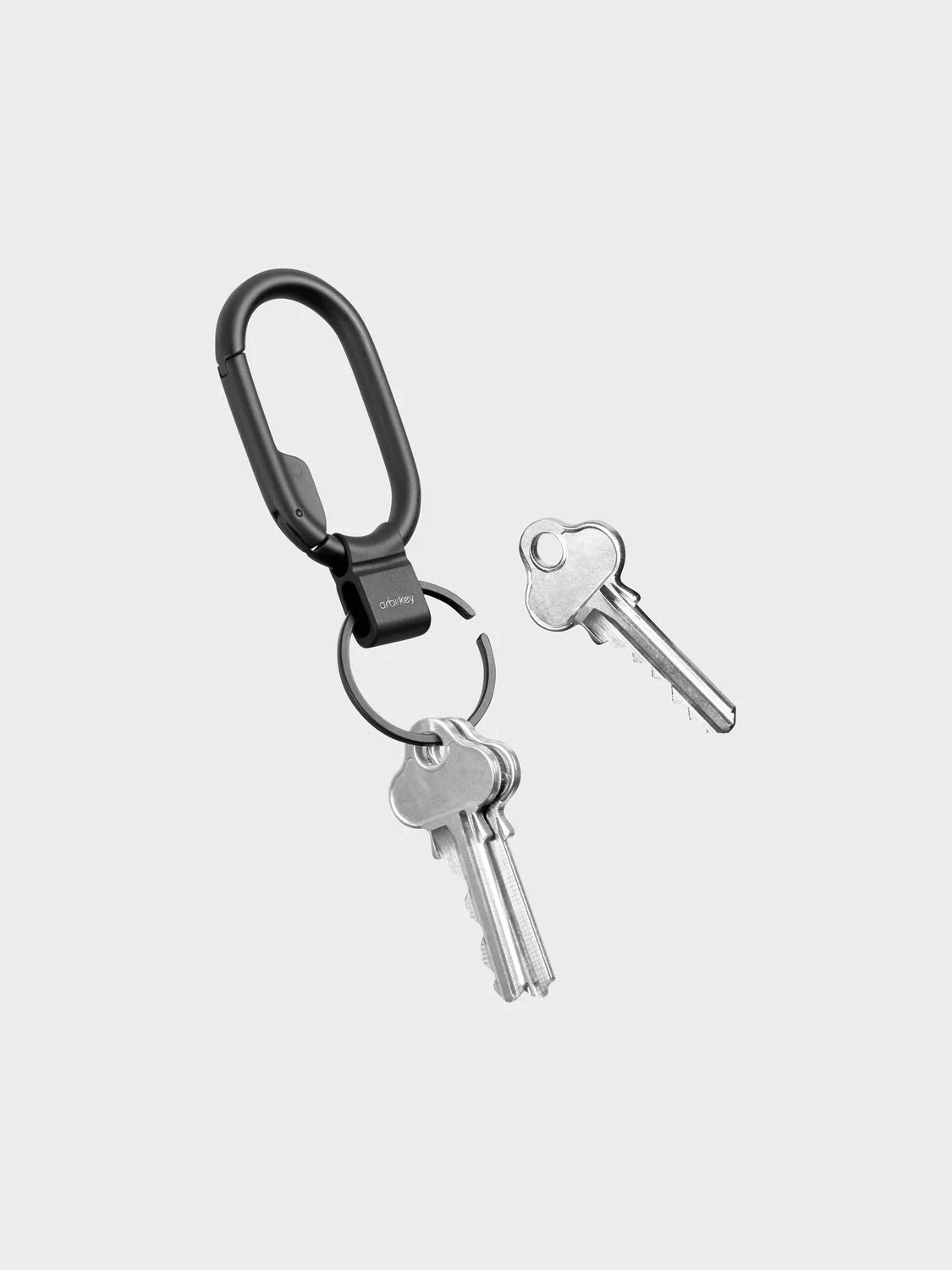 The Orbitkeys Clip Mini Keyring, a black minimalist carabiner, holds two silver keys on its ring while another silver key rests nearby, all arranged against a light gray background.