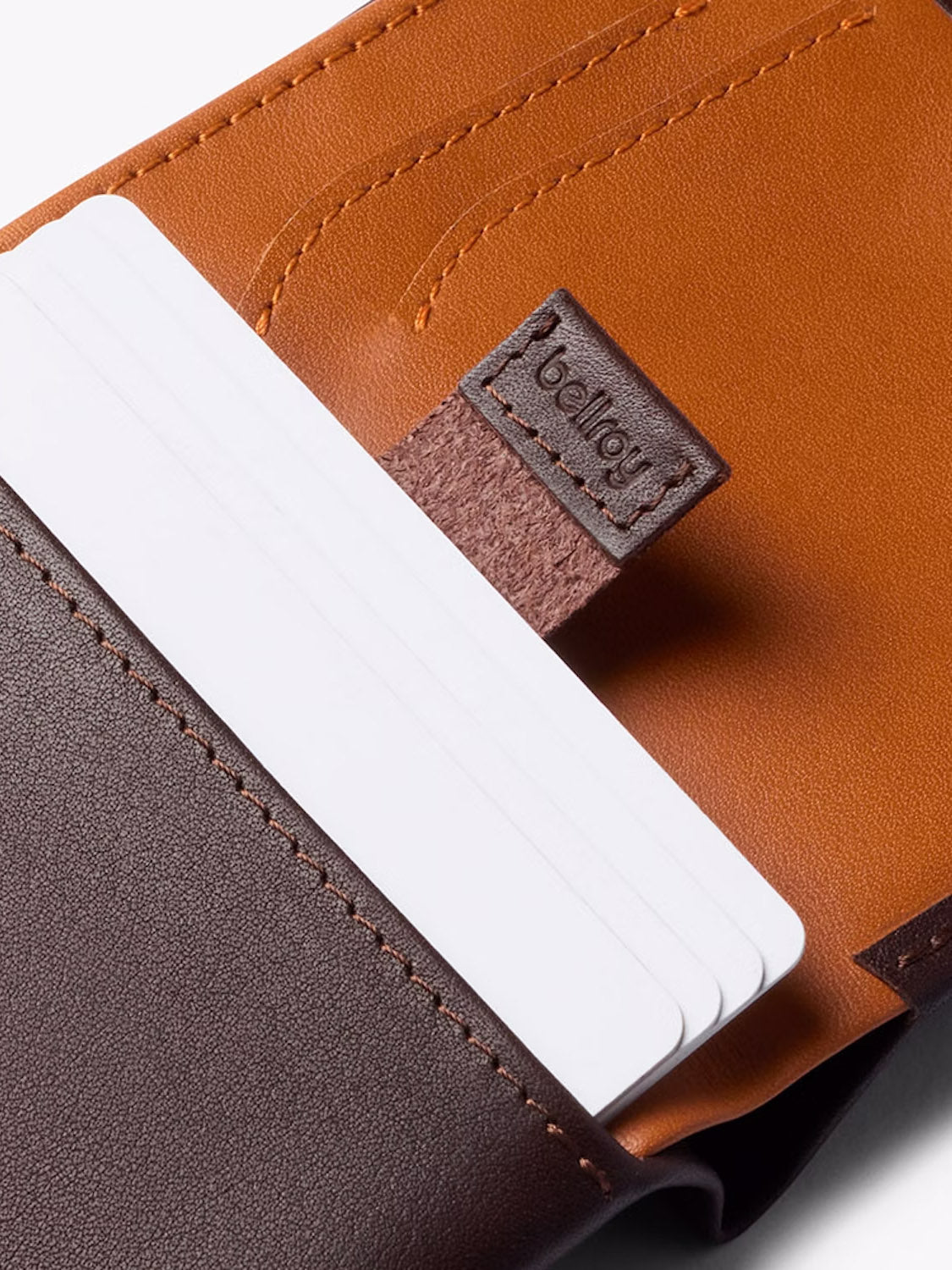 Close-up of a brown and tan Bellroy Note Sleeve Wallet with several white cards partially visible in an outer slot. The word "bellroy" is embossed on a leather tag inside the wallet.