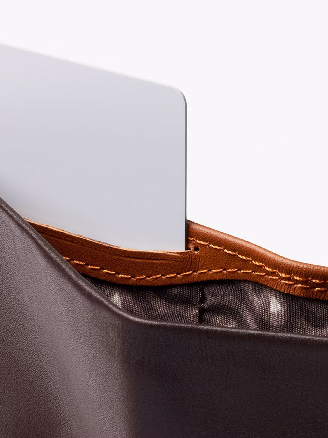A close-up of the Bellroy Note Sleeve Wallet in brown leather with orange stitching, partially open to reveal a white card, highlights its modern design and RFID protection against a plain light background.