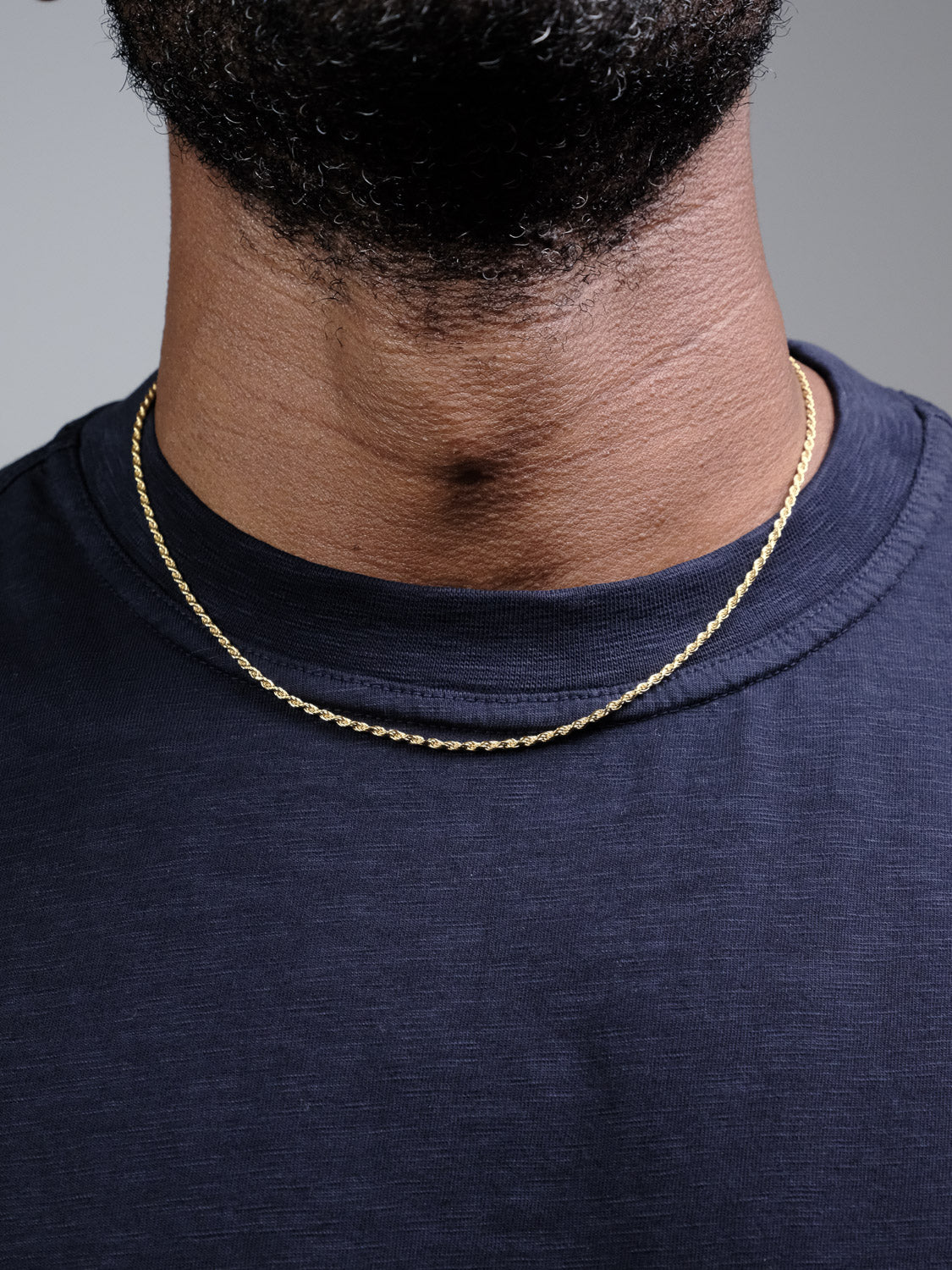 A close-up shows a person wearing the ID Menswear Orion 40 Rope Chain Necklace, a thin 3mm gold-plated chain, paired with a navy blue crewneck shirt. The lower face and beard are partially visible.