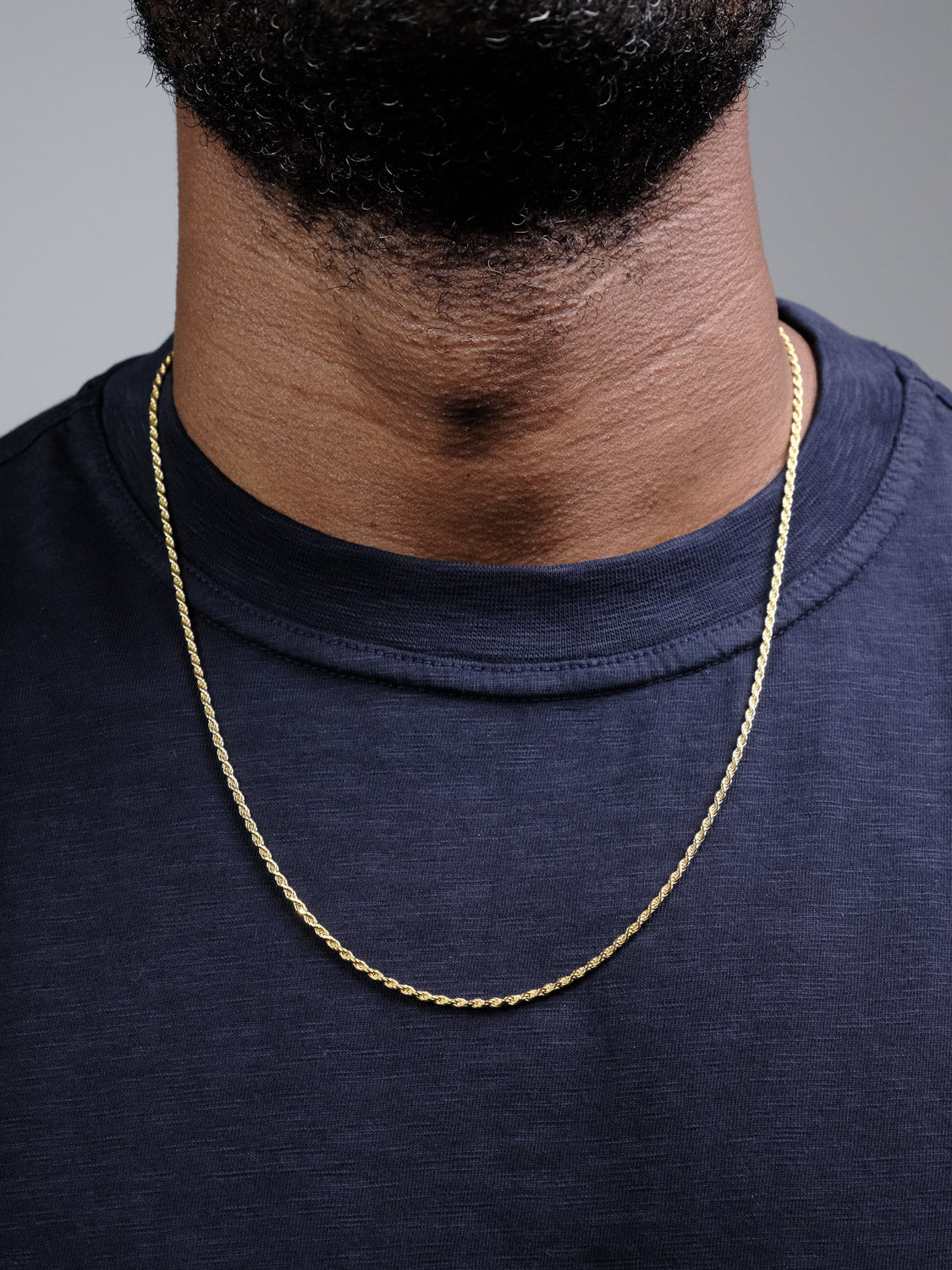Close-up of a person's neck and upper chest wearing a dark navy t-shirt and the Orion 40 Rope Chain Necklace by ID Menswear. A thin, gold-plated chain rests on their skin, with part of their beard visible above.