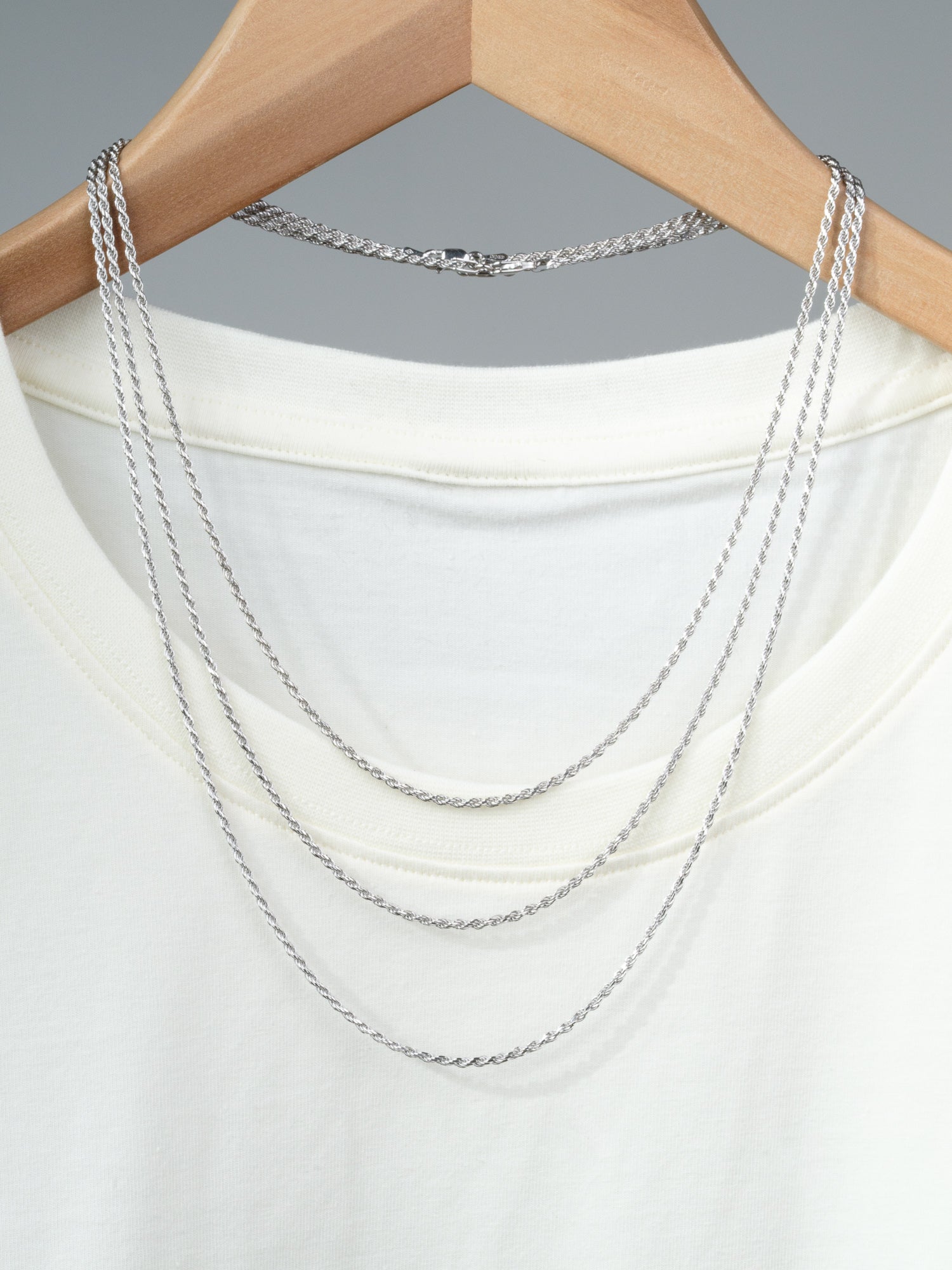 A white T-shirt on a wooden hanger with the Orion 40 Rope Chain Necklace by ID Menswear draped over the shoulders and neckline, displayed against a plain gray background.