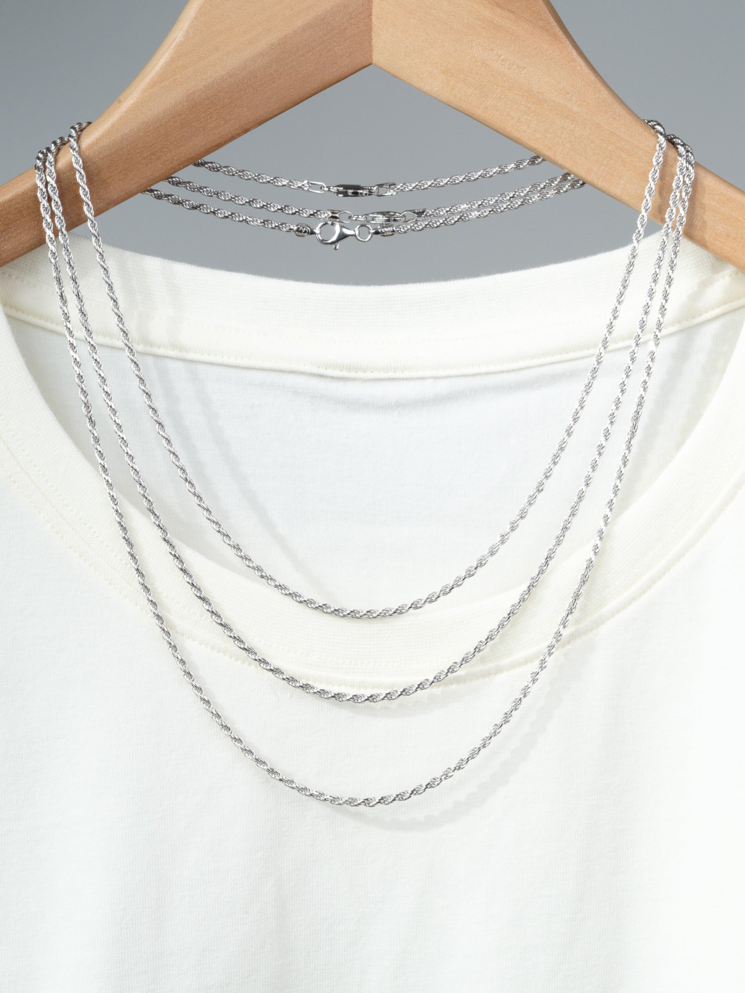 An Orion 50 Rope Chain Necklace from ID Menswear is draped over a white t-shirt on a wooden hanger, set against a plain gray background.