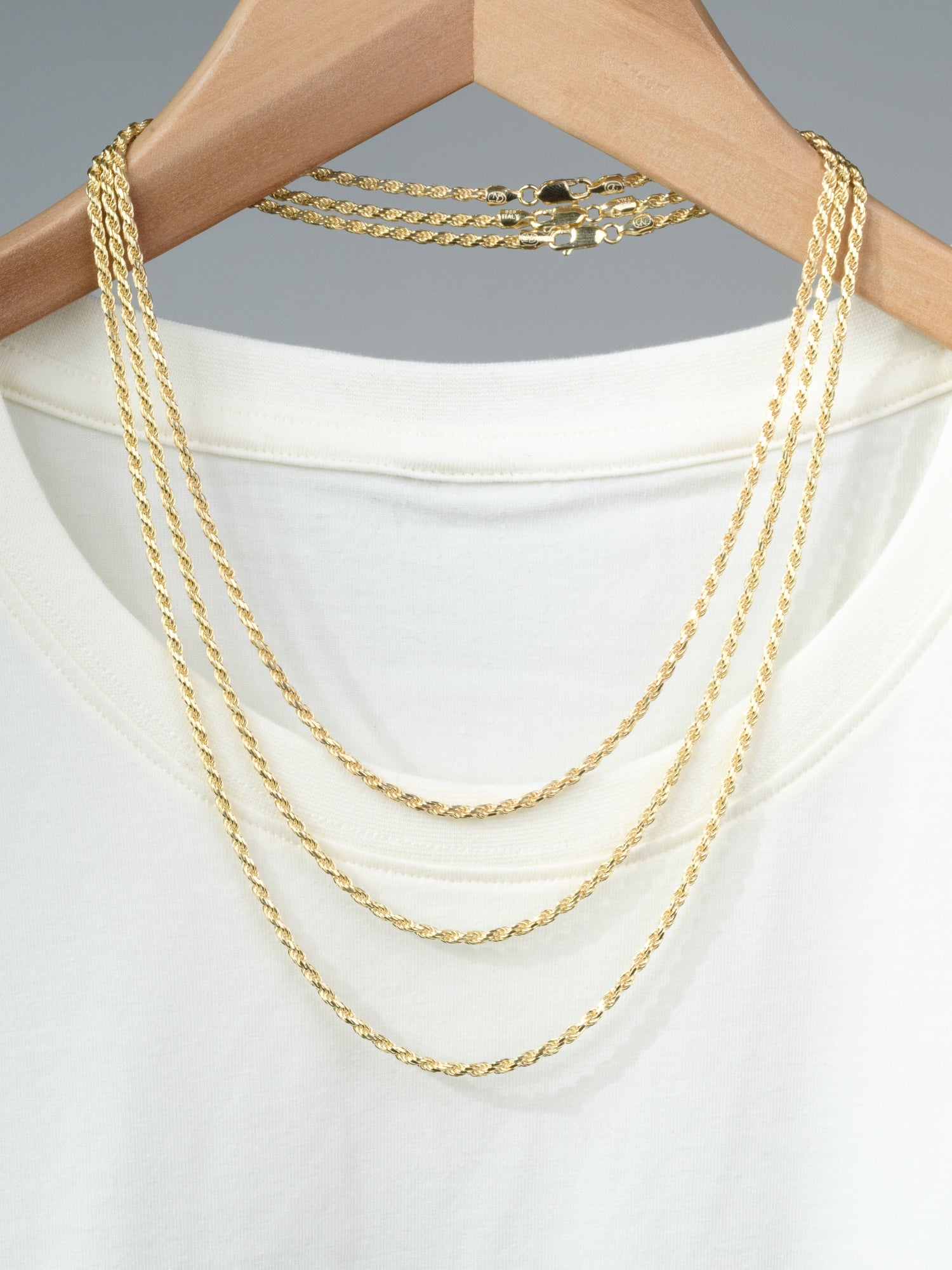 An Orion 60 Rope Chain Necklace by ID Menswear drapes over a white T-shirt on a wooden hanger, alongside two gold rope chains of varying lengths, all set against a neutral gray background.