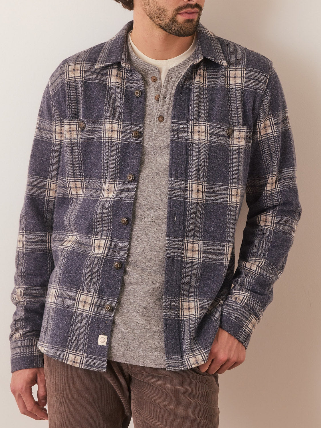 A man models the Marine Layer Pacific Stretch Twill Shirt—blue and beige plaid—layered over a gray henley with brown pants, standing with one hand in his pocket against a light-colored wall.