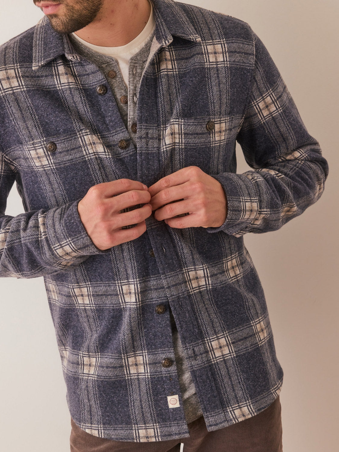 A person wearing the Marine Layer Pacific Stretch Twill Shirt in blue and beige plaid over a gray henley is buttoning up, with their face partially out of frame against a plain background.