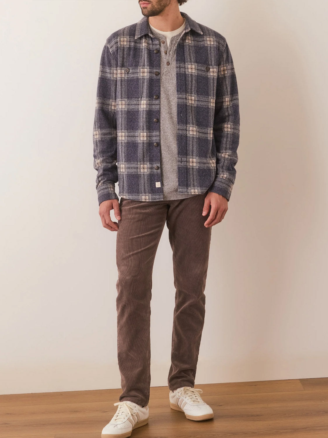 A man stands indoors wearing the Marine Layer Pacific Stretch Twill Shirt over a gray henley, paired with brown corduroy pants and white sneakers. His hands are by his sides, and his face is partially out of frame.