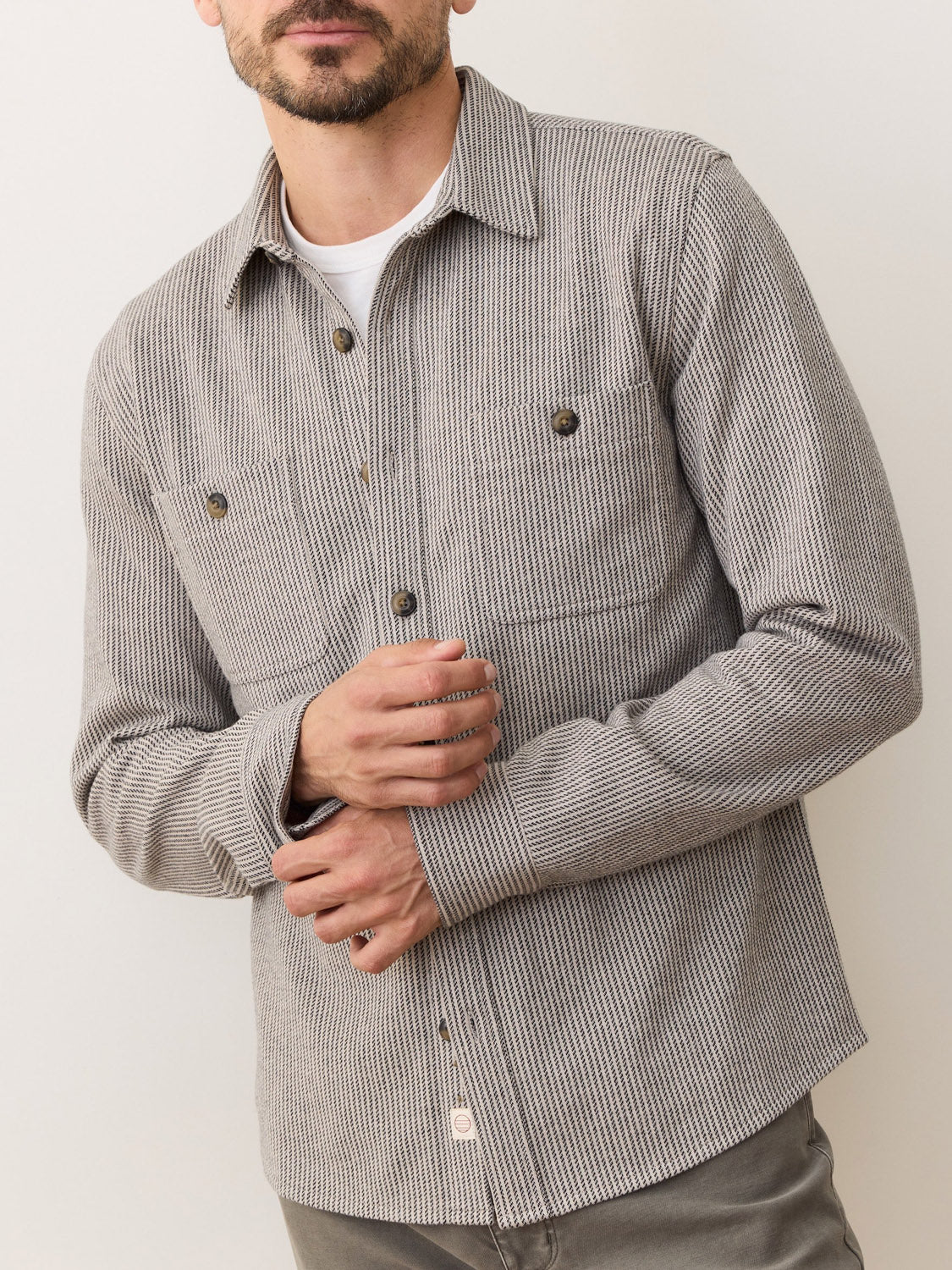 A man wears the Marine Layer Pacific Stretch Grey Twill Overshirt over a white t-shirt with grey pants, posing with hands clasped. The classic fit and medium-weight fabric provide style and comfort against a neutral background.