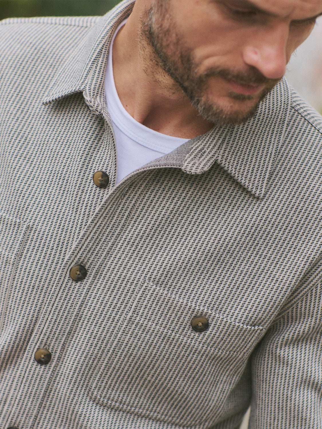 A man wears the Marine Layer Pacific Stretch Grey Twill Overshirt over a white tee, looking down and away. The overshirt features a subtle pattern, two chest pockets, and dark buttons.