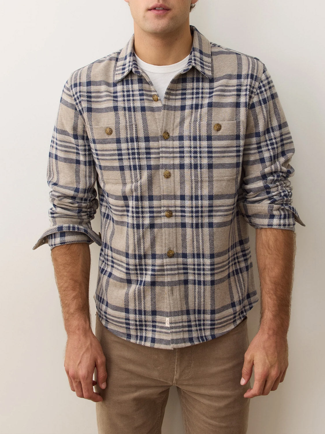 A man stands against a plain background in the Marine Layer Pacific Stretch Plaid Twill Overshirt, beige and blue, worn over a white undershirt with rolled-up sleeves and light brown pants. His face is partially out of frame.