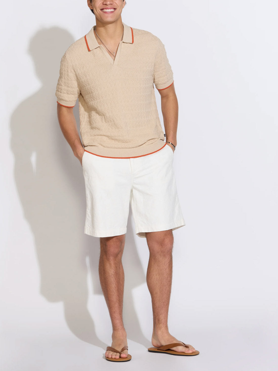 A smiling person stands against a white background wearing the Civil Society Palani Johnny Collar Sweater Polo in beige with orange trim, paired with white shorts and brown sandals. Their hands are in their pockets.