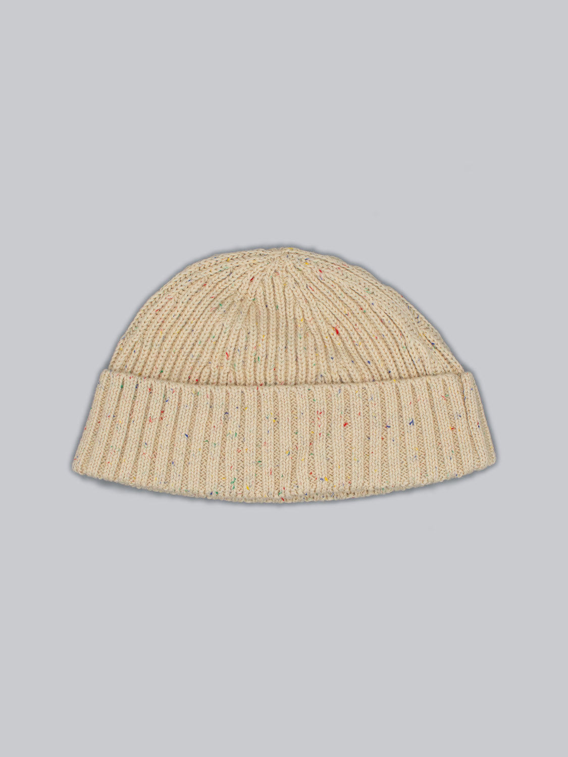 The Paro Speckled Cotton Fishermans Beanie by ID Menswear is a beige knit hat with a folded cuff, crafted from speckled yarn and featuring multicolored speckles. Its laid flat against a light gray background, highlighting its timeless design.