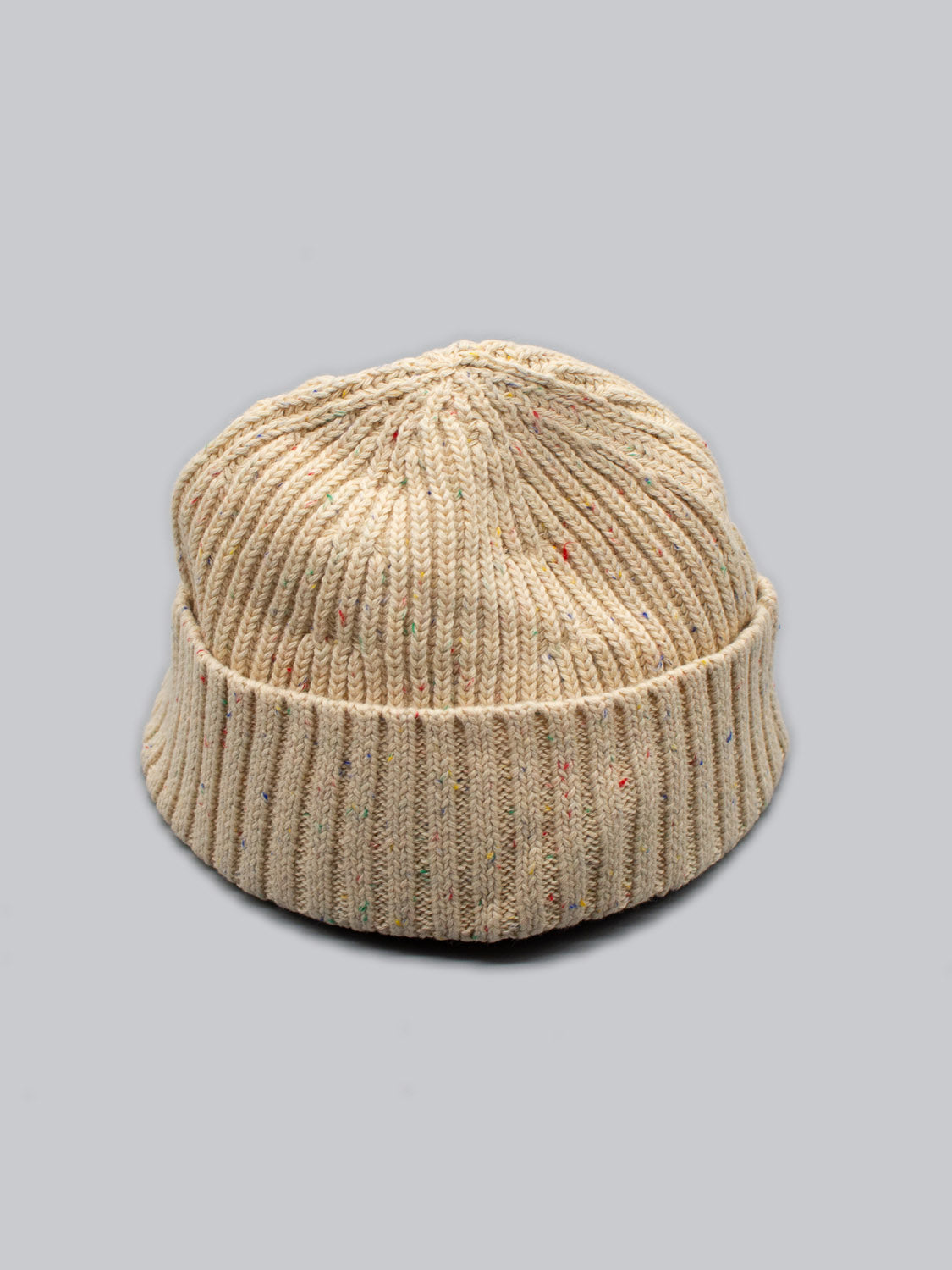 The Paro Speckled Cotton Fishermans Beanie by ID Menswear boasts a timeless design with a folded brim, colorful speckles woven into beige knitwork, all set against a plain gray background.