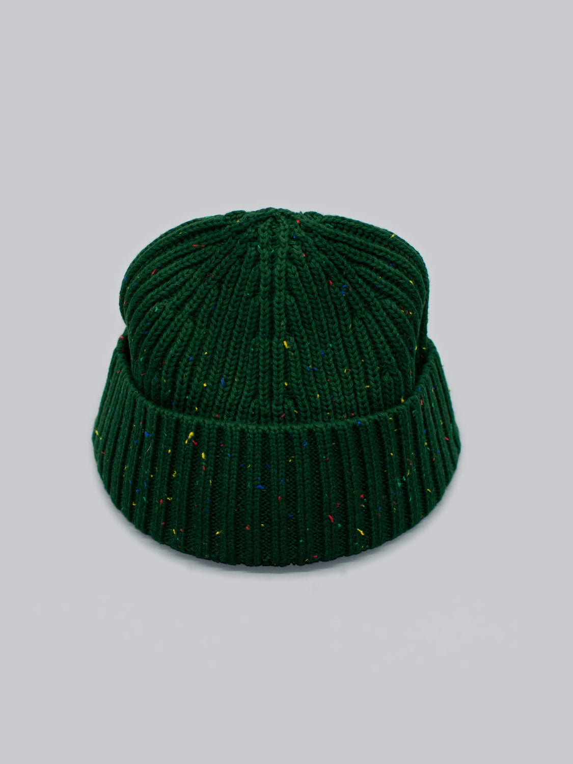 The Paro Speckled Cotton Fishermans Beanie by ID Menswear in dark green features a timeless design with colorful speckles woven into the speckled yarn, set against a light gray background, making it stylish and versatile.