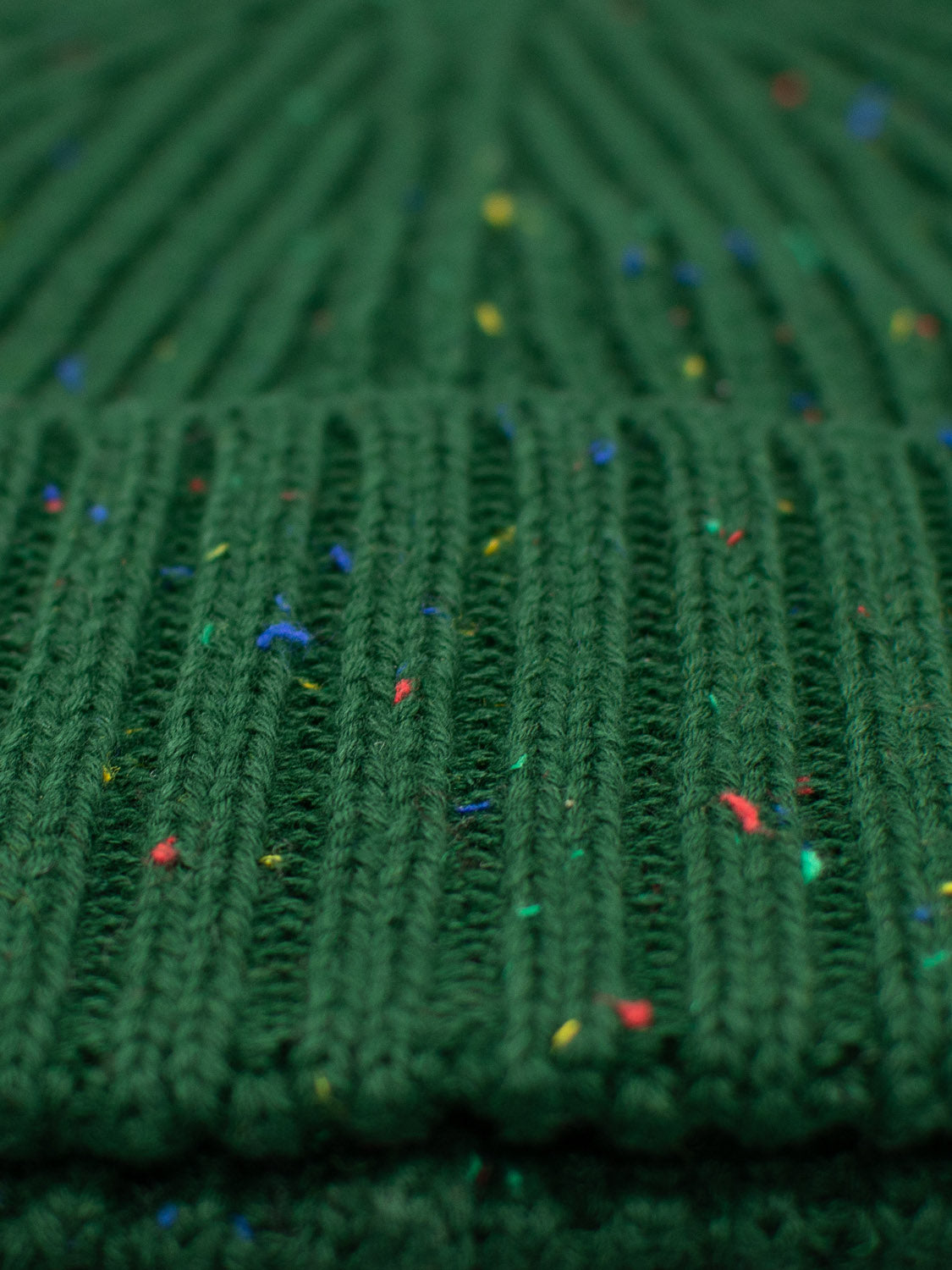 A close-up of a knitted green fabric with a ribbed texture and speckled yarns in blue, red, and yellow echoes the timeless design of the Paro Speckled Cotton Fishermans Beanie by ID Menswear, seamlessly blending style and color.
