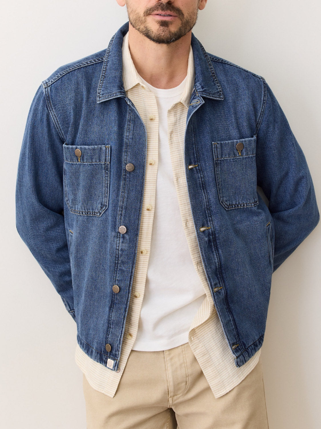 A man stands against a plain light background with his hands in his pockets, wearing the Marine Layer Patrick Denim Overshirt over a cream shirt and white tee, paired with beige pants. His face is partially visible.