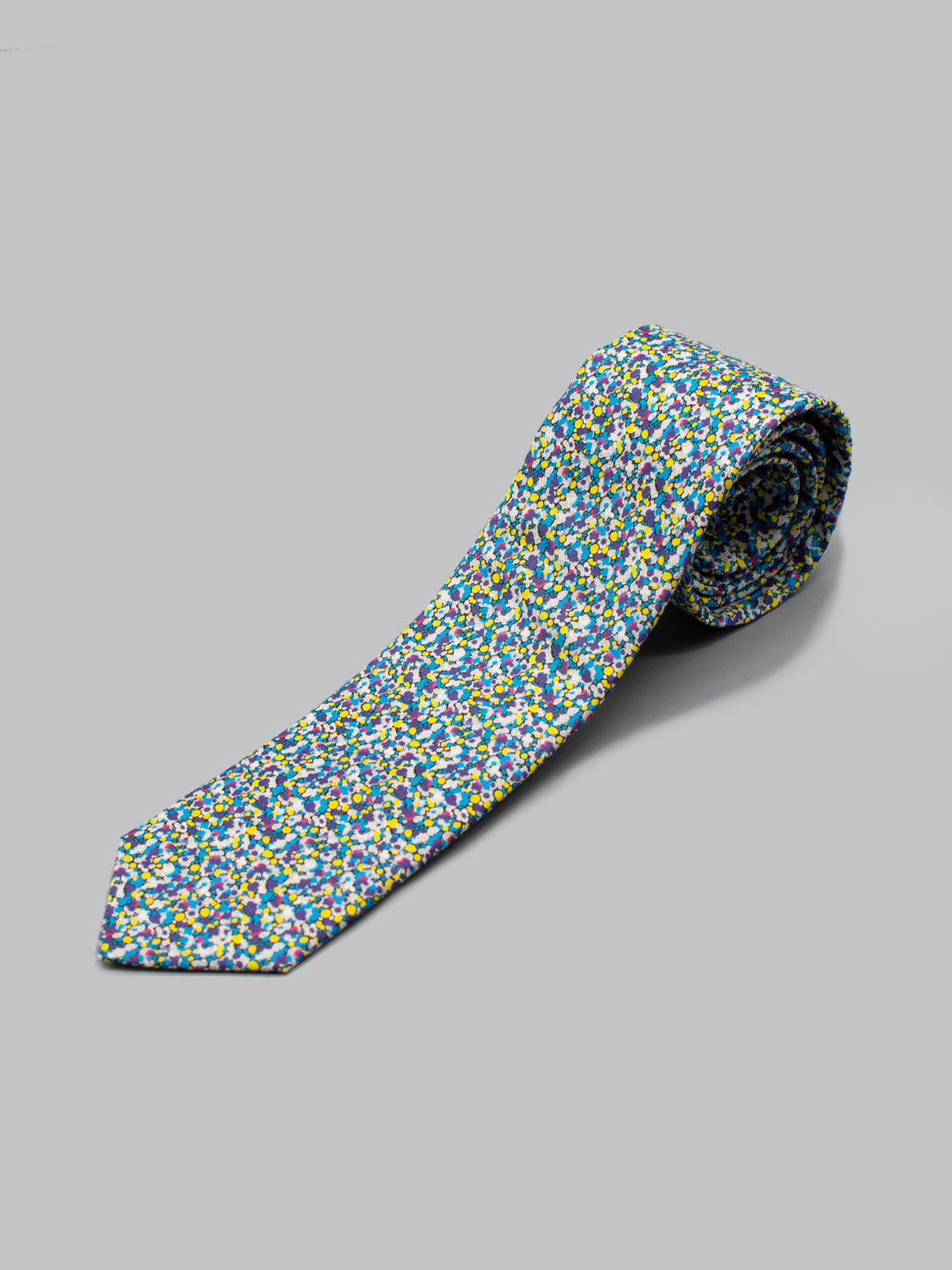 The Liberty of London Tana Lawn™ Pepper by ID Menswear is a rolled-up necktie handmade in Brooklyn, featuring a colorful abstract pattern of blue, yellow, green, and white spots on a light gray background.