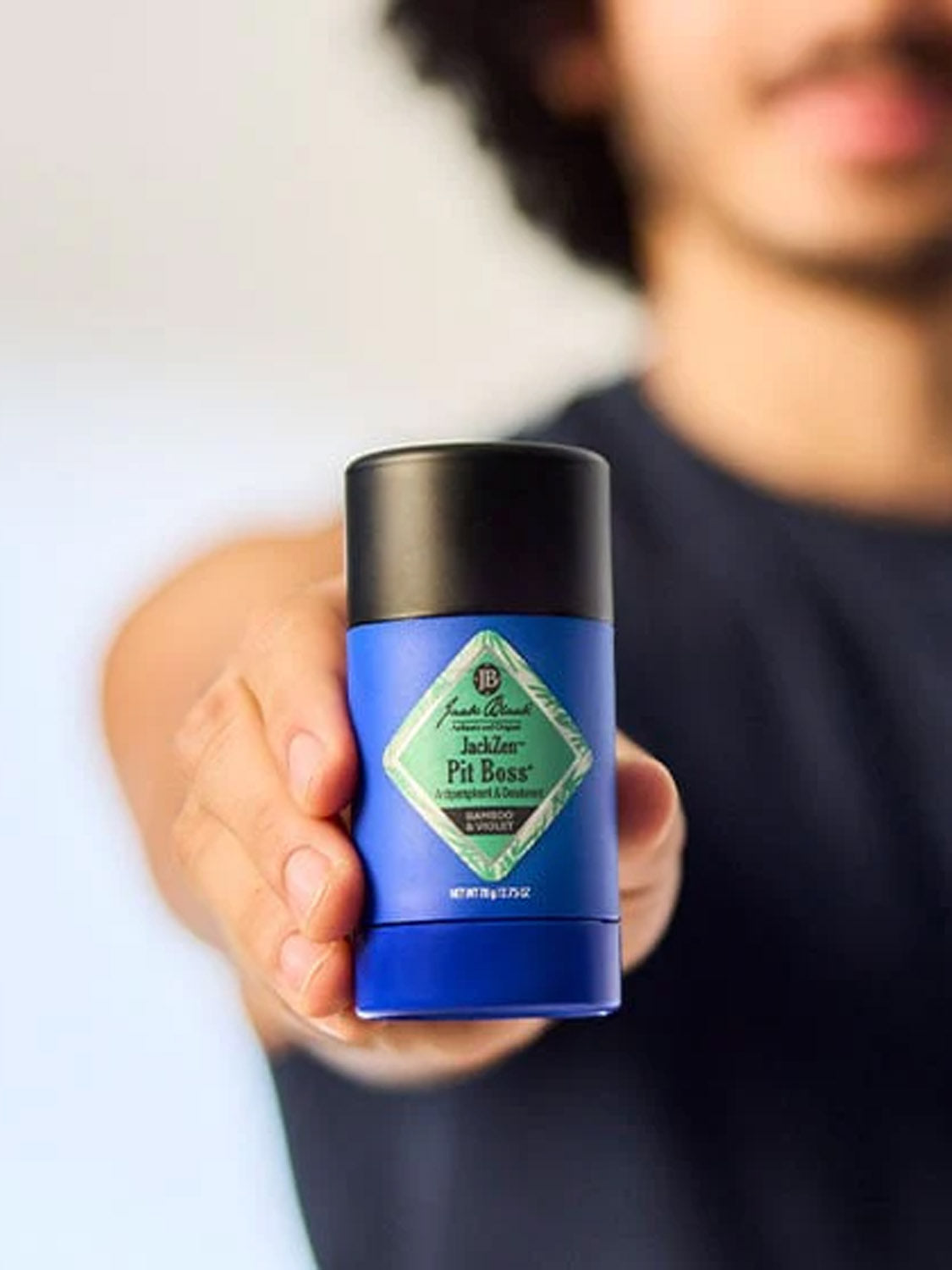 A person holds a blue stick of Jack Black JackZen™ Pit Boss® Antiperspirant & Deodorant toward the camera, with the label visible and their face blurred in the background, emphasizing its long-lasting odor protection.
