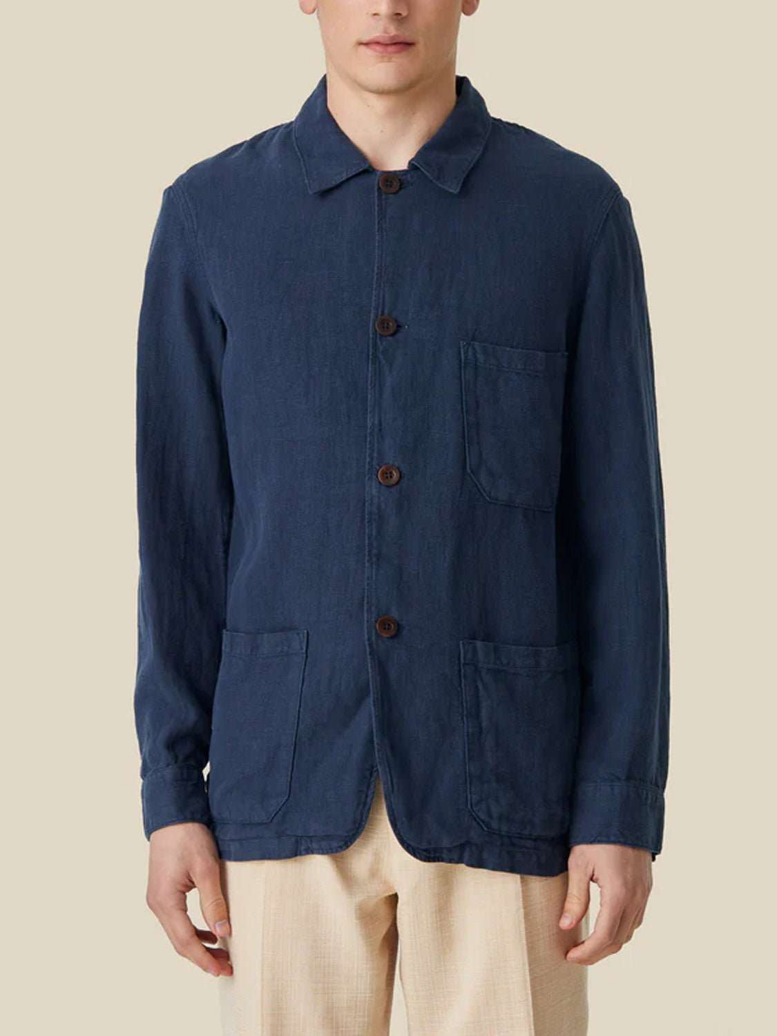 Wearing the Portuguese Flannel Labura Linen Jacket, a dark blue button-up with three front pockets, highlights its Portuguese heritage. Its perfectly paired with light beige pants on a neutral backdrop.