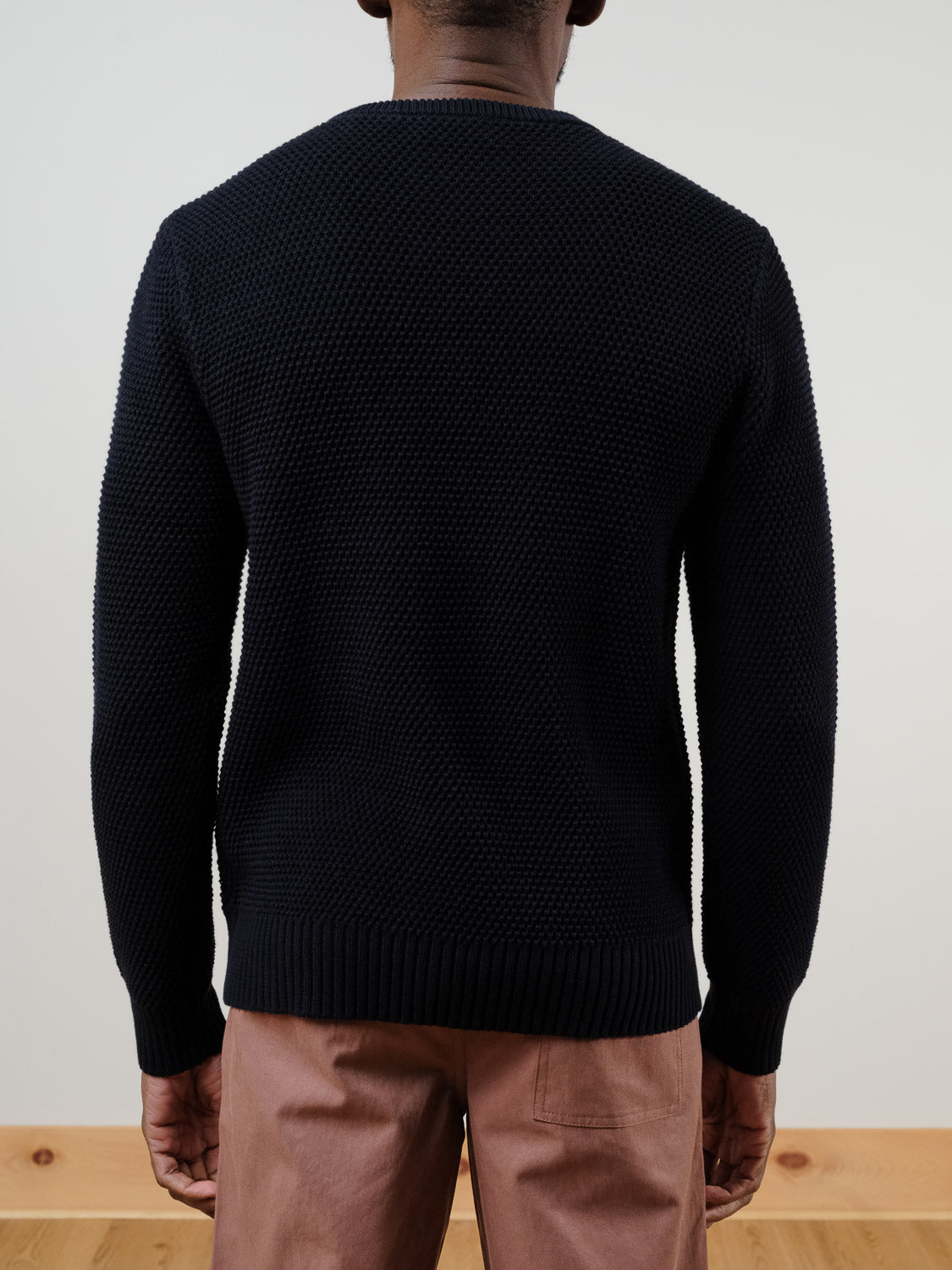 A person faces away from the camera, wearing the Quebec Chunky Knit Sweater by ID Menswear in textured dark navy, paired with light brown pants, set against a plain light background and wooden flooring.