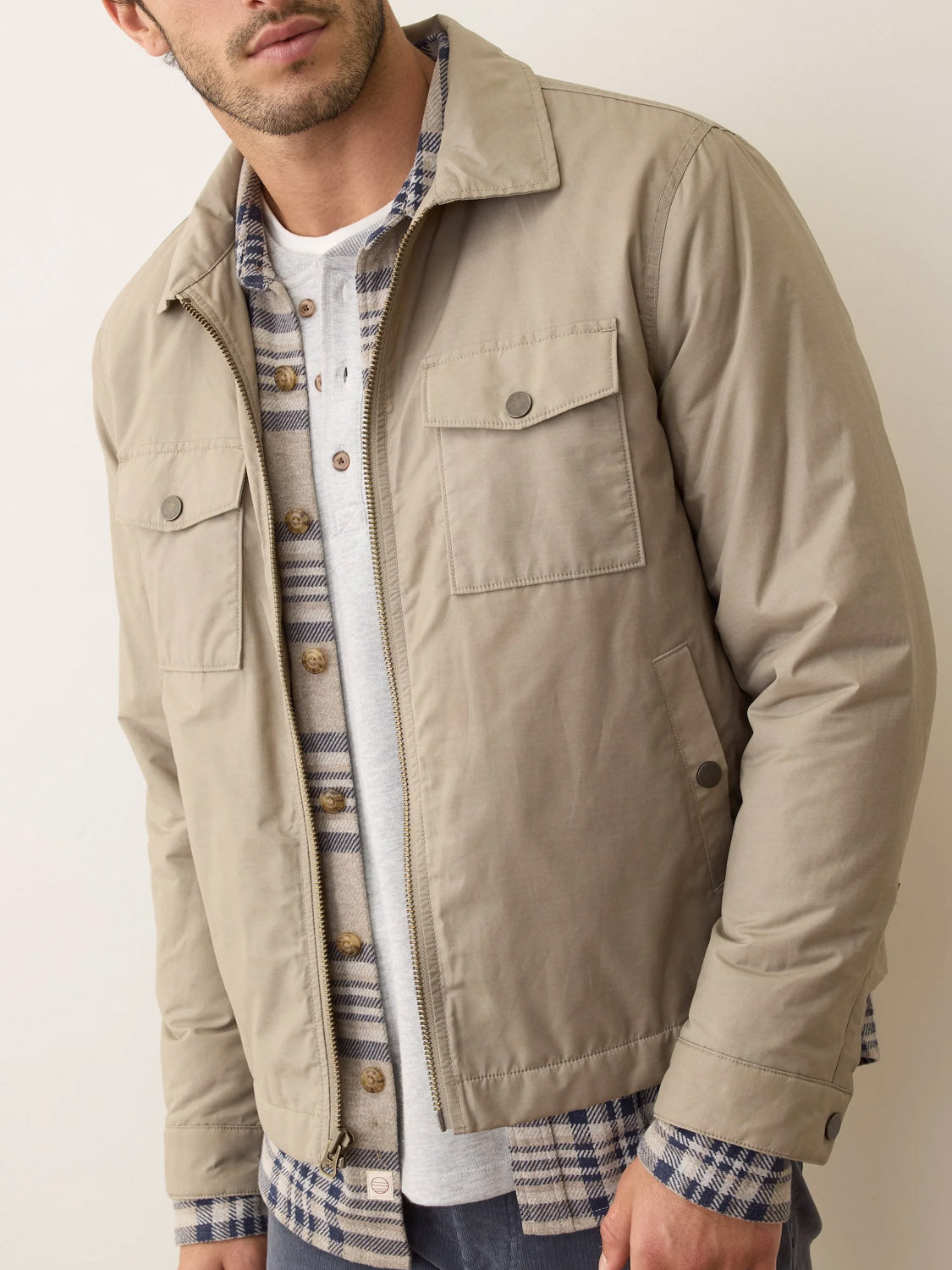 A man stands against a plain light background wearing Marine Layer’s Reynolds Dry Wax Trucker Jacket in light beige over a blue and white plaid shirt and a light gray henley. His face is partially visible, showing short dark hair and light facial hair.
