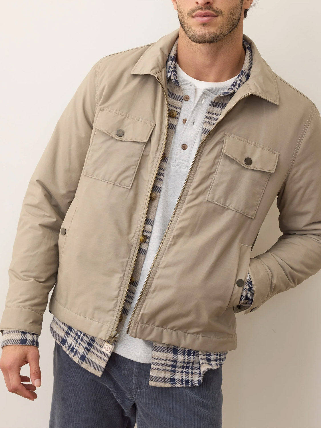 A man stands against a plain light wall, one hand in his pocket, wearing the Marine Layer Reynolds Dry Wax Trucker Jacket over a gray henley and blue plaid shirt. His face is partially out of frame.