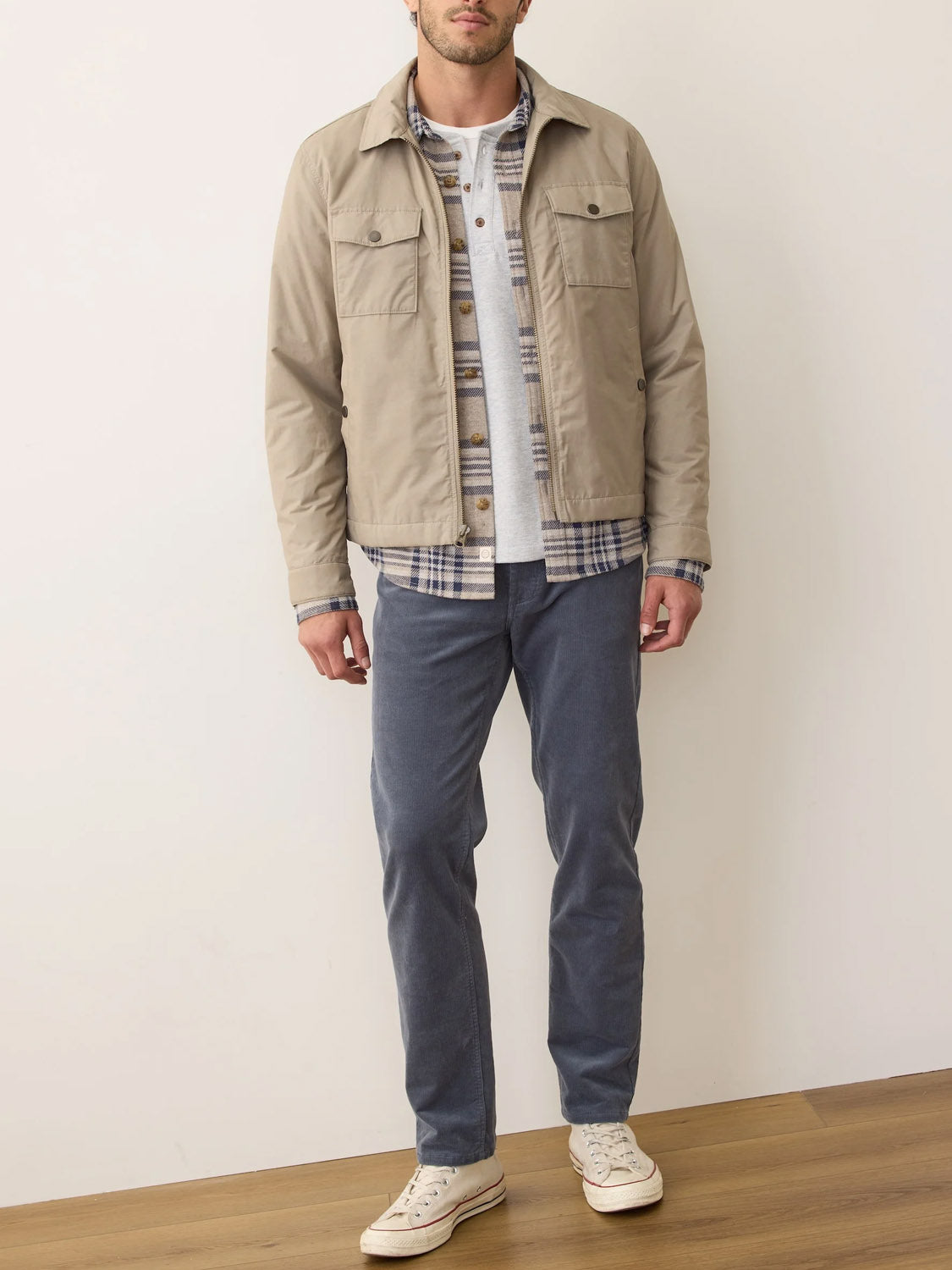 A man stands indoors on a wooden floor wearing the Marine Layer Reynolds Dry Wax Trucker Jacket over a plaid shirt and gray tee, paired with blue pants and white sneakers against a plain background.