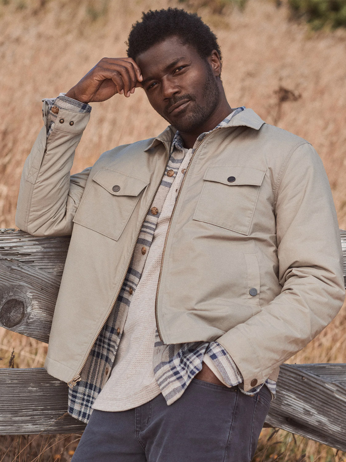 A man with short curly hair leans on a wooden fence in a field, wearing the Marine Layer Reynolds Dry Wax Trucker Jacket over layered shirts and dark pants. He gazes thoughtfully at the camera, with tall grass in the background.