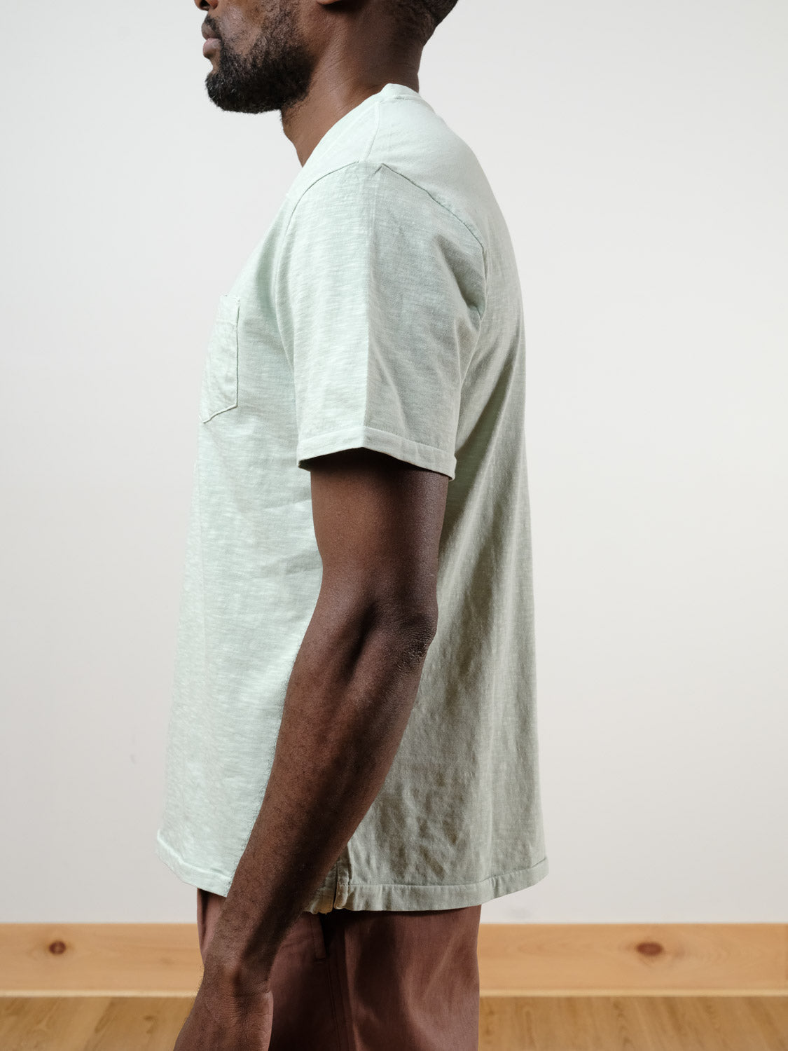 A man stands sideways wearing the Rogue Henley, a light green short-sleeve 100% cotton t-shirt by ID Menswear, with brown pants against a plain light-colored wall and wooden floor.