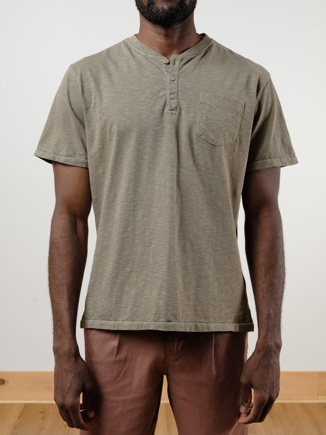 A person wears the olive green Rogue Henley by ID Menswear, crafted from 100% cotton slub yarn with a chest pocket, paired with brown pants. They stand against a plain light background; their face is not visible.