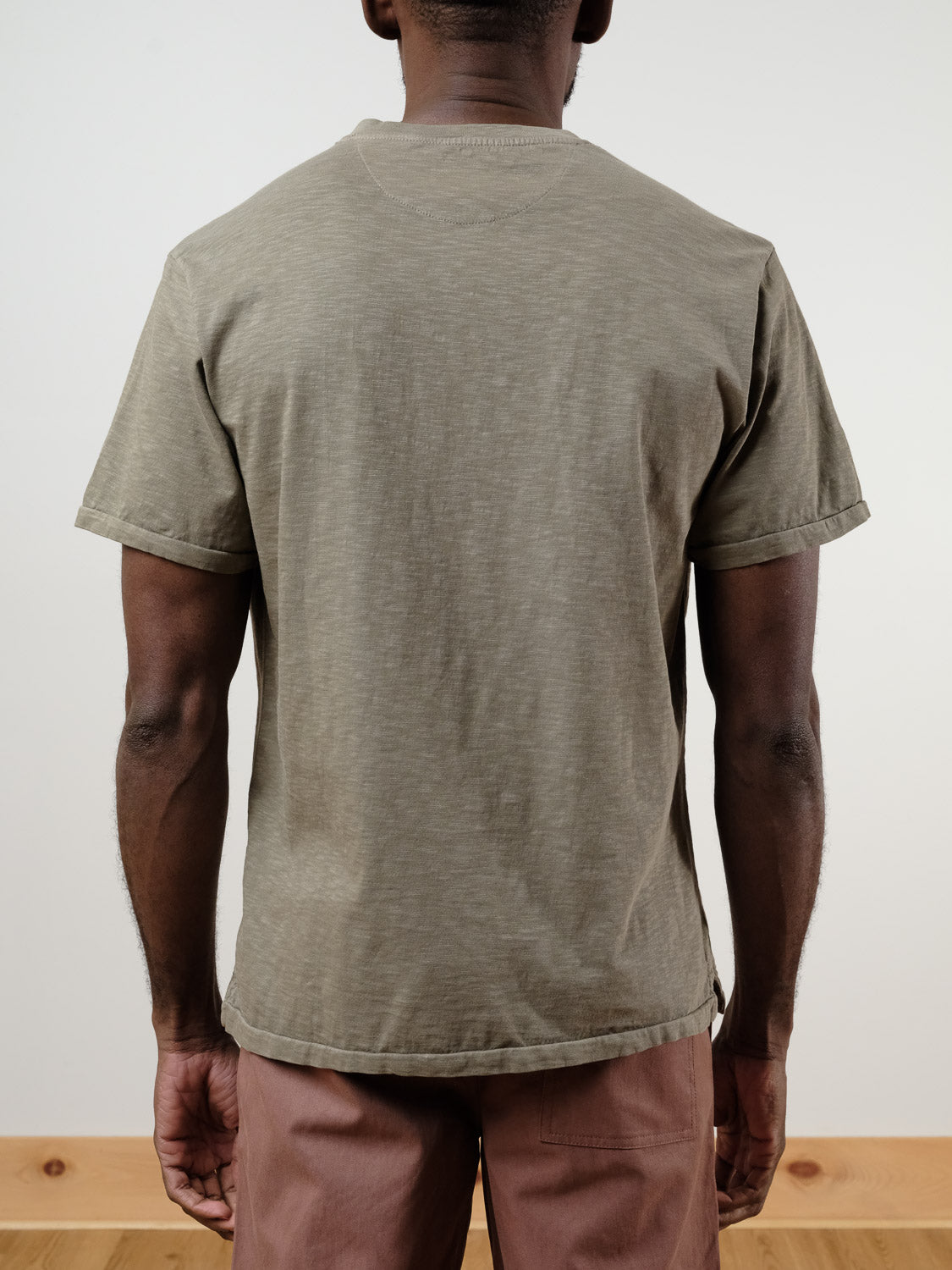 A person stands indoors facing away, wearing the ID Menswear Rogue Henley—a short-sleeve, light olive-green 100% cotton t-shirt—with light brown pants. The plain background features light walls and a wooden floor trim.