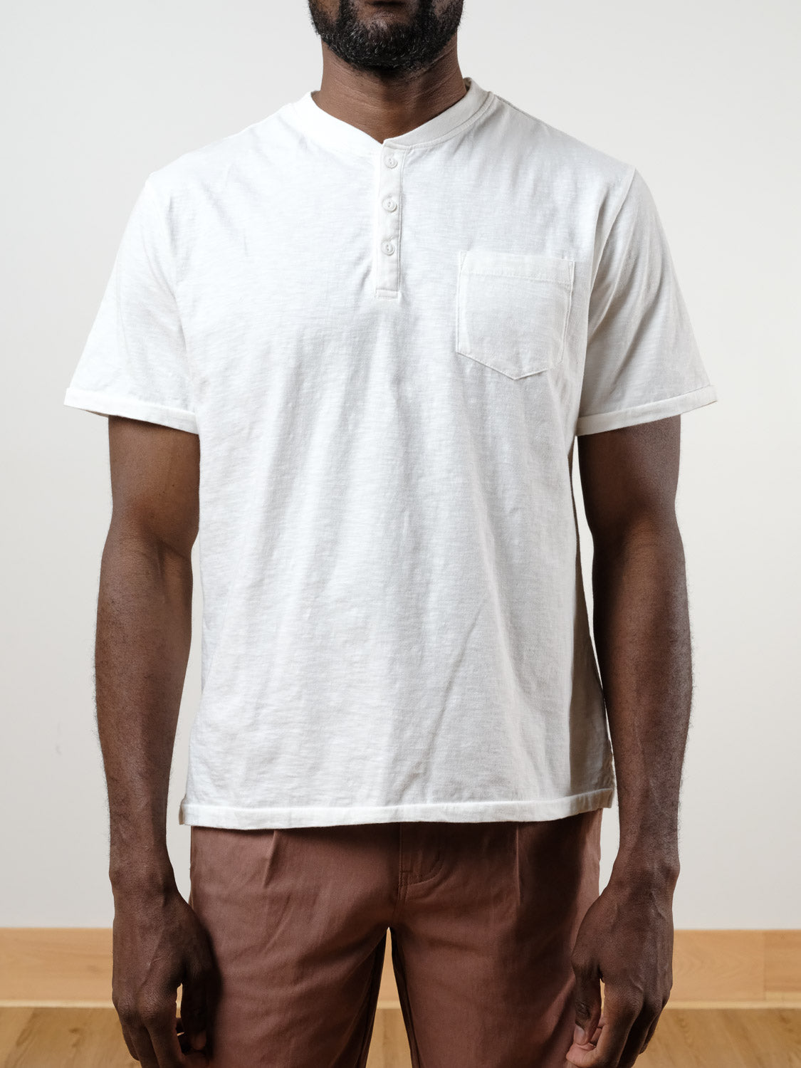 A man stands against a plain background wearing the ID Menswear Rogue Henley, crafted from 100% cotton slub yarn with a chest pocket, paired with brown pants.