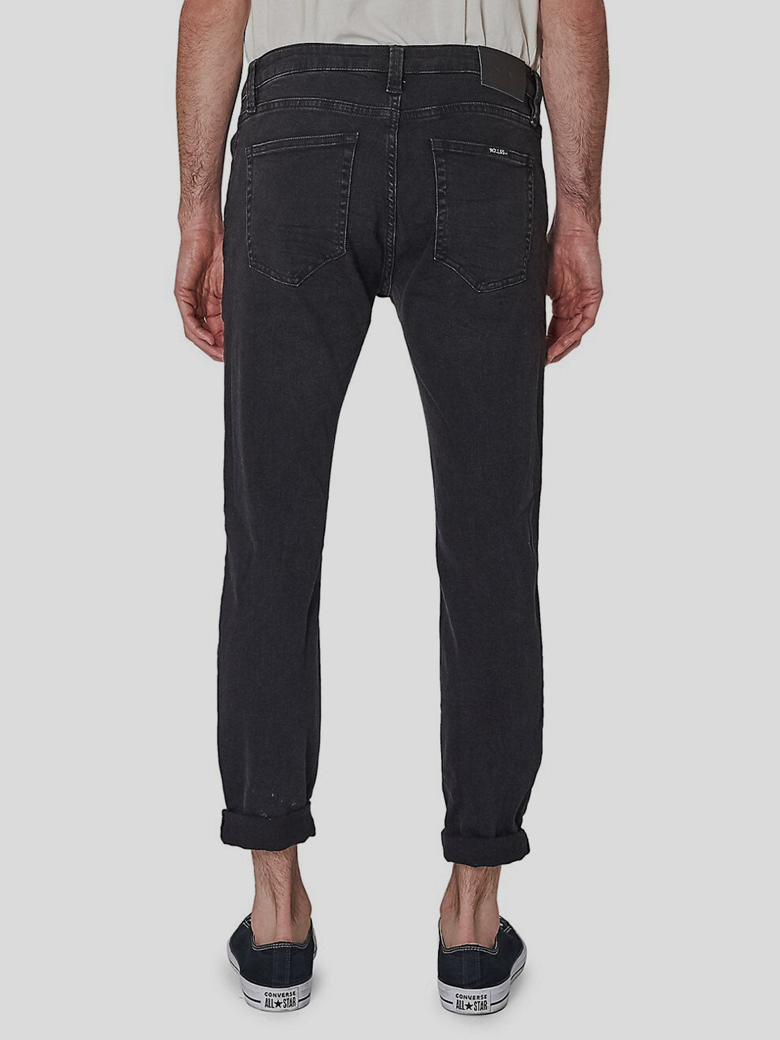 Someone wearing Rollas Rollies Slim Fit Jean, styled with rolled cuffs and black sneakers, stands with their back to the camera, showcasing the jeans' rear view against a plain light gray background.
