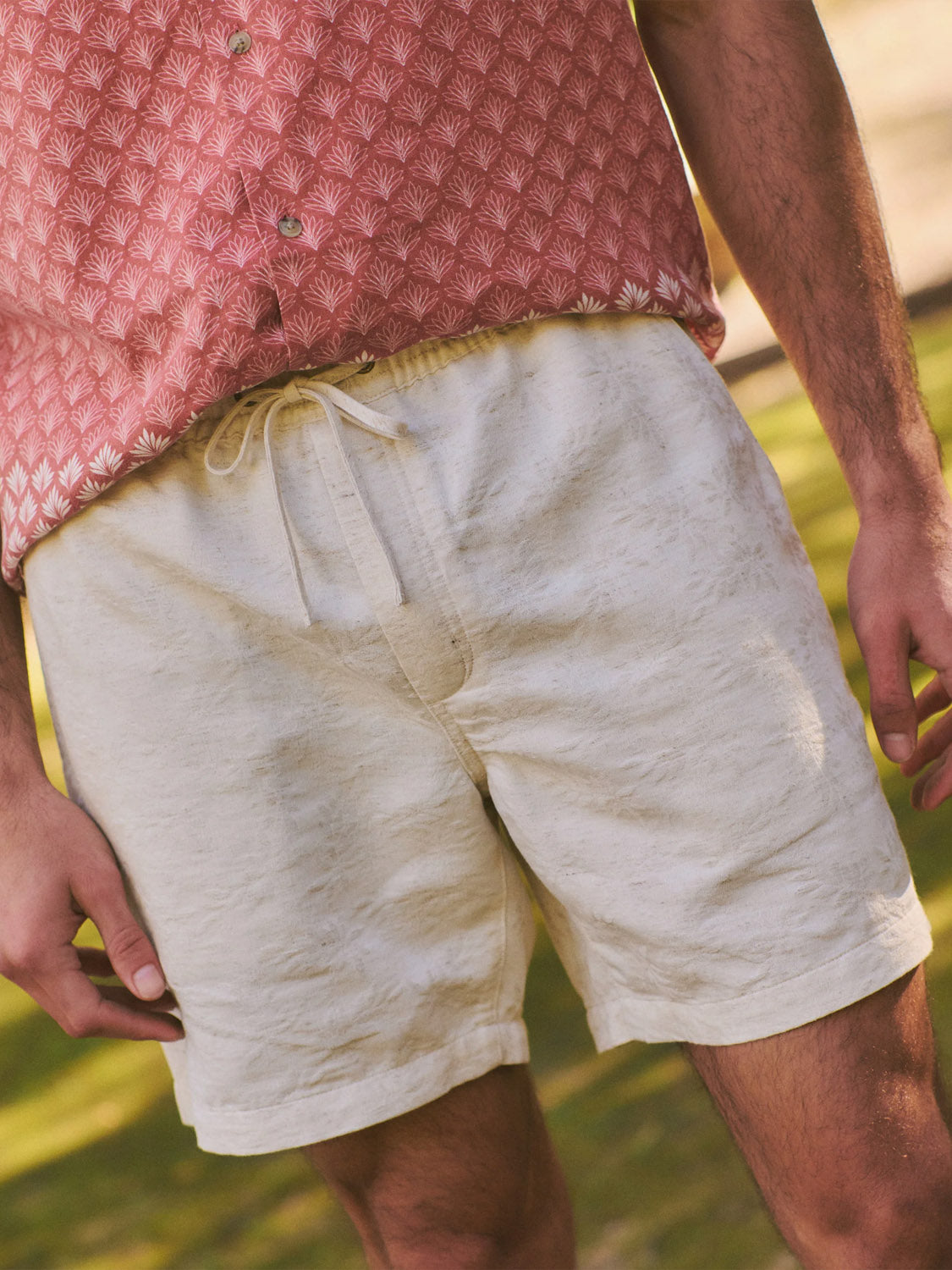 Wearing Marine Layer’s Saturday Beach Short 6" in light beige jacquard, a person stands relaxed outdoors on sunlit grass, paired with a patterned pink resort shirt.