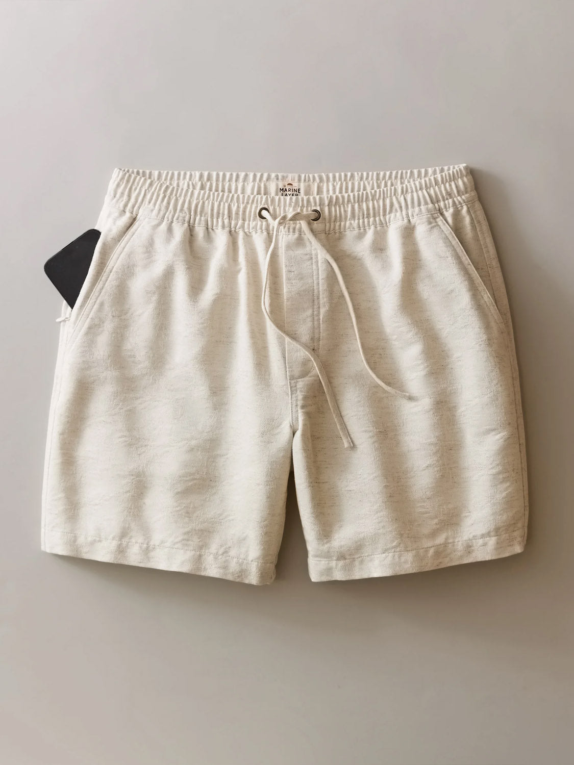 The Marine Layer Saturday Beach Short 6" in beige jacquard features an elastic waistband, side pockets, and a small black item peeking from the left pocket, showcased flat on a neutral background.