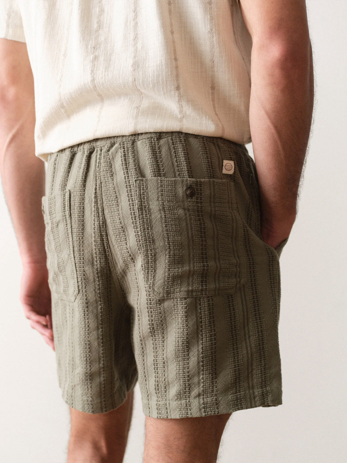 Model shown from waist down and back wearing Marine Layer Saturday Dobby Short 6" in textured olive green with back pockets, made from 100% cotton Dobby weave, paired with a cream short-sleeve shirt. One hand is in their pocket.