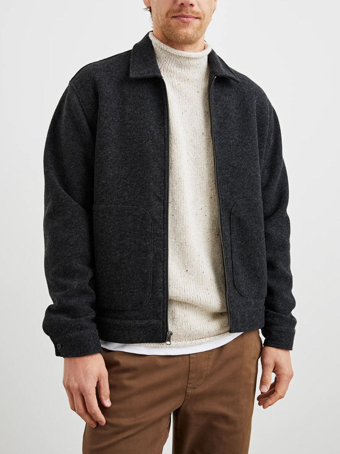 A man models the Rails Shael Jacket, a dark gray zip-up winter outerwear piece, layered over a cream knit sweater and brown pants. He stands against a plain white background with his face partially visible.