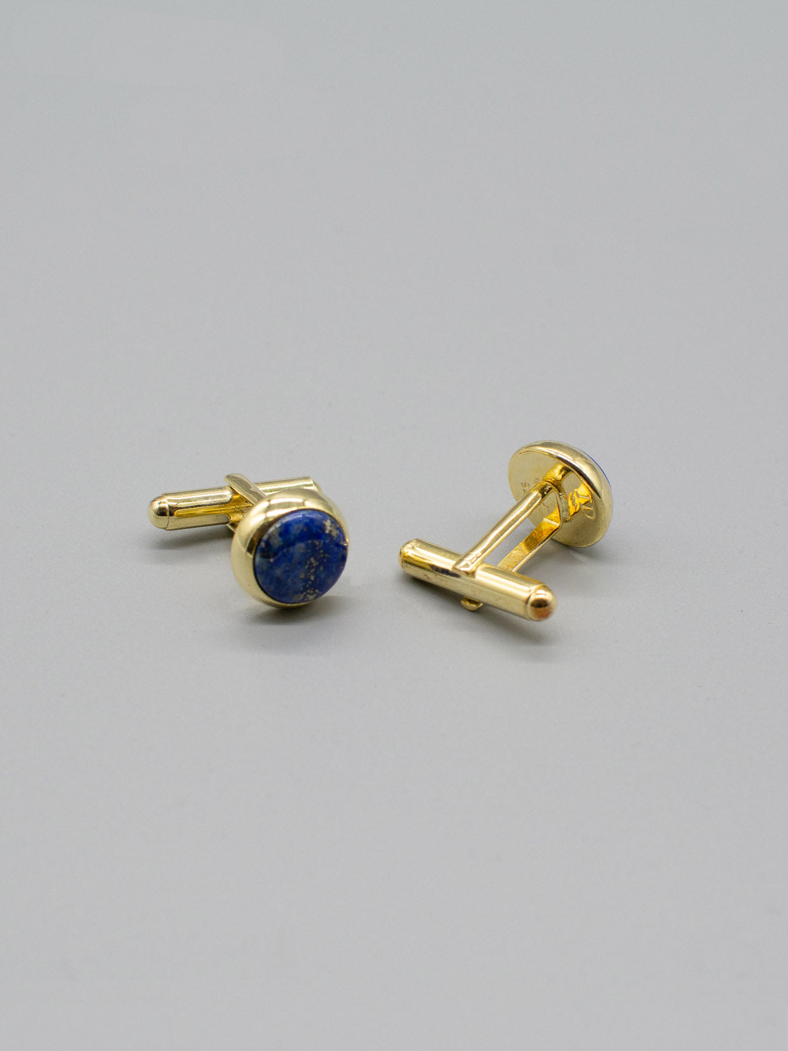 Two Gold Plated Silver Gemstone Cufflinks by ID Menswear are on a plain gray surface. One is upright showcasing its bullet back mechanism, while the other is flat, highlighting the gemstones deep blue hue.