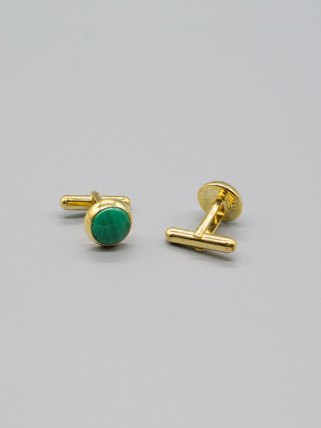 Two exquisite ID Menswears Gold Plated Silver Gemstone Cufflinks feature smooth round green stones in a refined inlay. Positioned on a light gray surface, one cufflink faces up while the other displays its intricate back design.