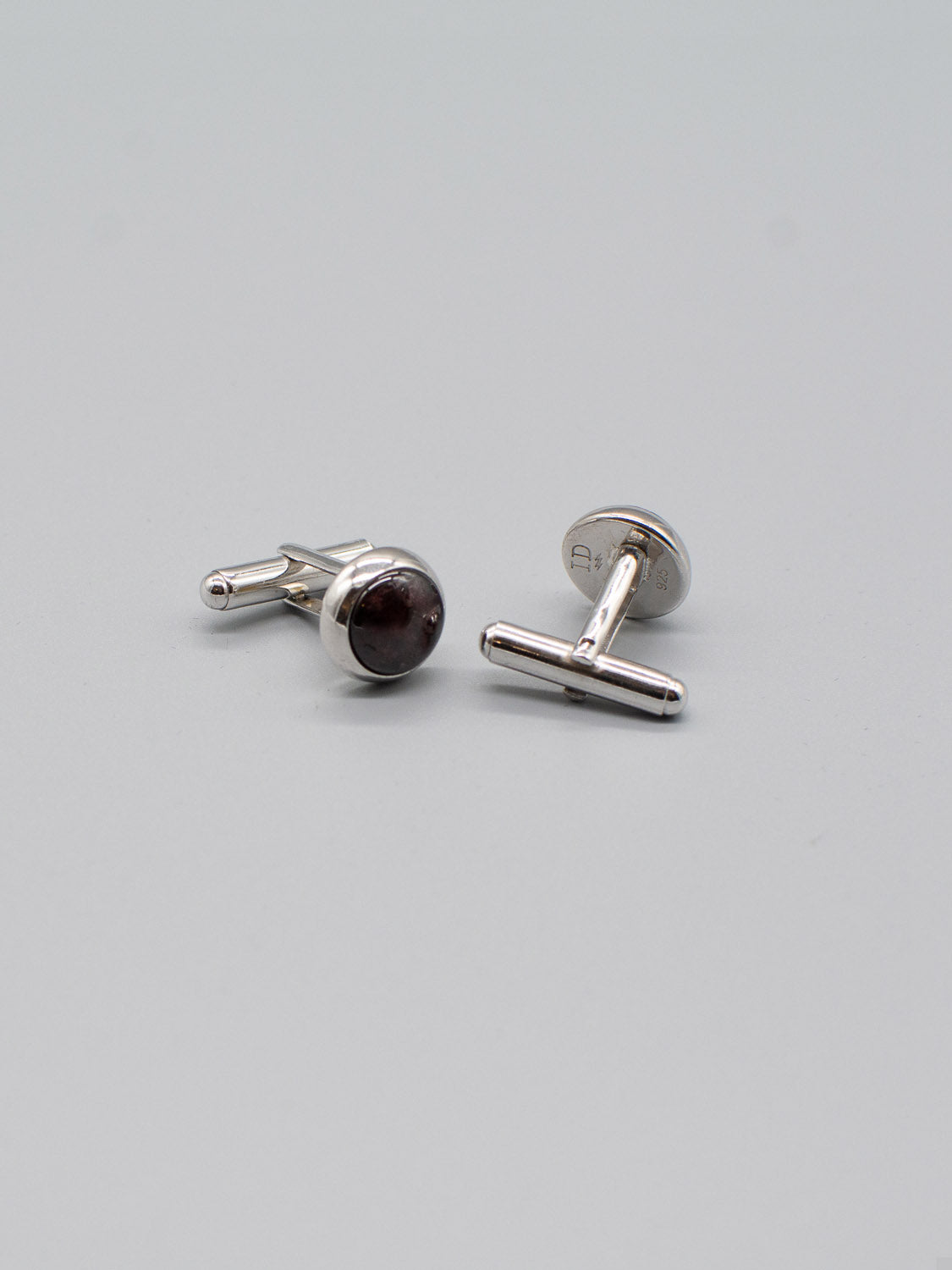 Two ID Menswear Rhodium Plated Silver Gemstone Cufflinks are on a plain gray surface, featuring round, dark stones. One stands upright showing off its genuine gemstone inlay, while the other lays flat to reveal its toggle closure.