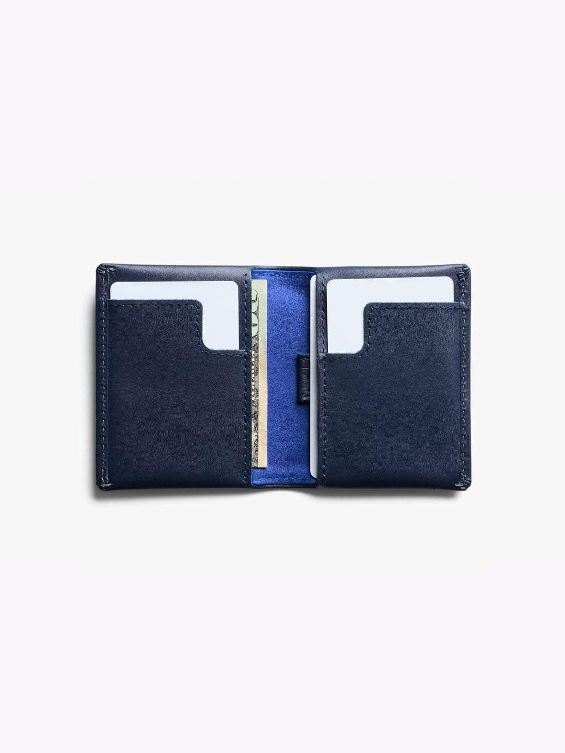 The Bellroy Slim Sleeve Wallet in navy blue leather is open, revealing two card slots with white cards and a central pocket holding a folded bill. Its interior features a contrasting blue lining.